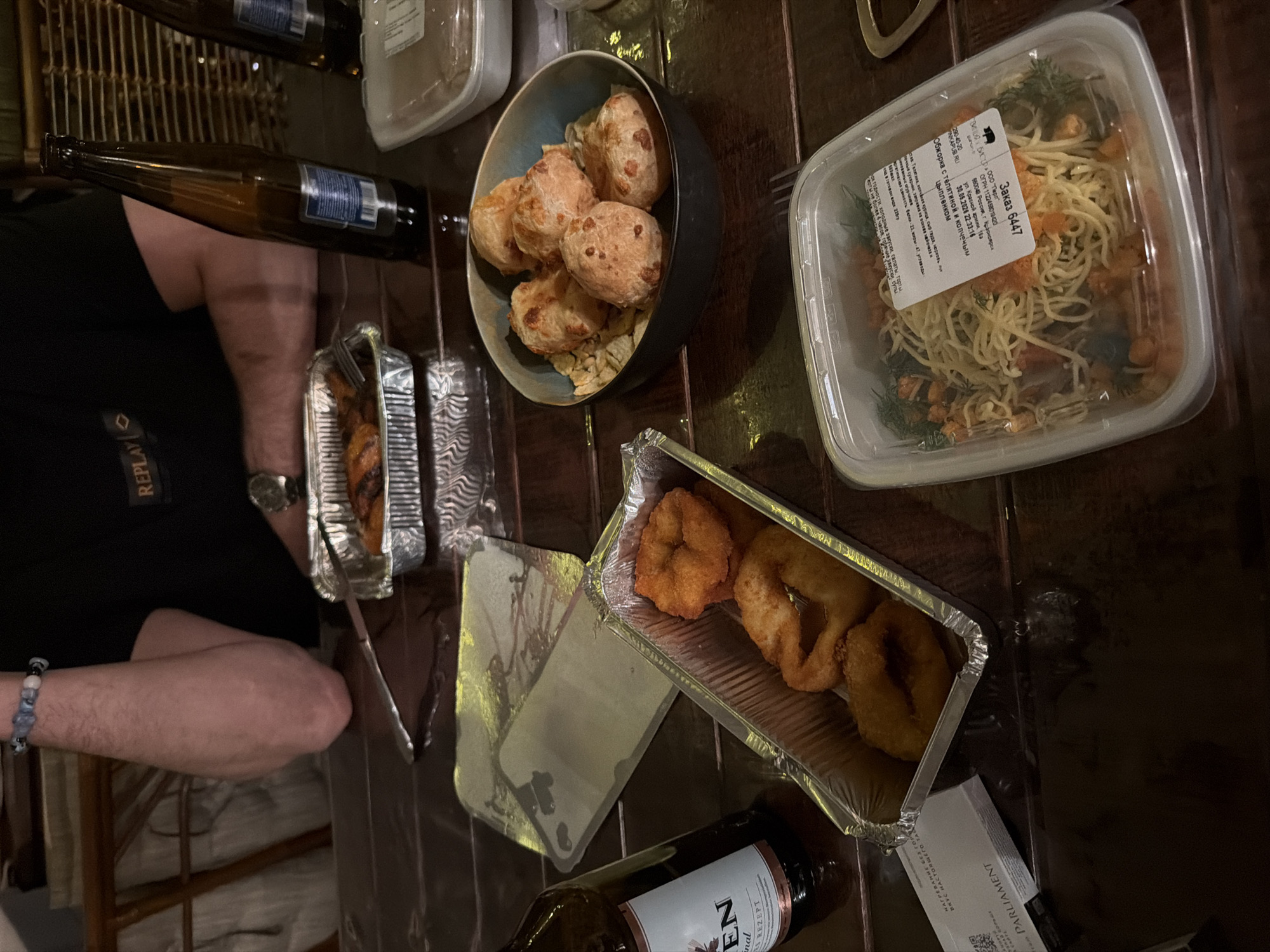The image depicts a table with several food containers and bottles, seemingly a meal in progress.
There is a person visible on the left side of the image wearing a black shirt with "REPLAY" visible. The person has a wrist with a bracelet.
Various food items are present, including what appears to be pasta with salmon in a clear container, several fried items in an aluminum container, and round pastries in a black bowl. There are also several unopened food containers, bottles of alcoholic beverages, and what may be cigarette packaging on the table.
A label on one of the containers reveals that the food order is number 6447, from "Обжорка с телятиной и копченым цыпленком" in Krasnoyarsk, Russia, and the company is "ГВМНЫЙ БИСЕР ООО "Перл" " on ul. Krasnoy army, 6a. The order was placed at 22:33:16 on 30.05.2026.
There is also a packaging labeled "PARLIAMENT."