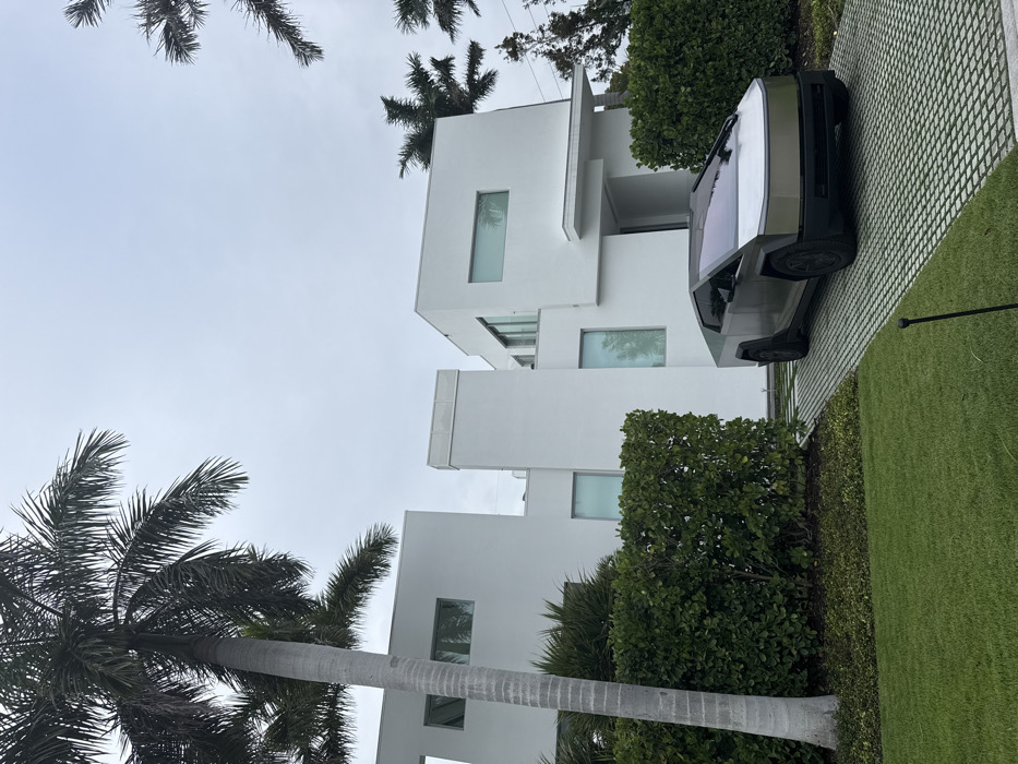 The image displays an exterior scene featuring a modern residential property in Naples, Florida, United States of America. A grey and silver Tesla Cybertruck is parked diagonally on a light-colored grid paver driveway, positioned in front of a multi-level, contemporary white stucco house. The residence exhibits sharp angles, flat roofs, and numerous rectangular windows, some appearing tinted or reflective. Green, manicured hedges line the perimeter of the property and the base of the house, separating it from a vibrant green lawn in the foreground. Several tall palm trees with grey trunks and green fronds are prominent features, framing the house against an overcast, light grey sky. No individuals are present in the scene.