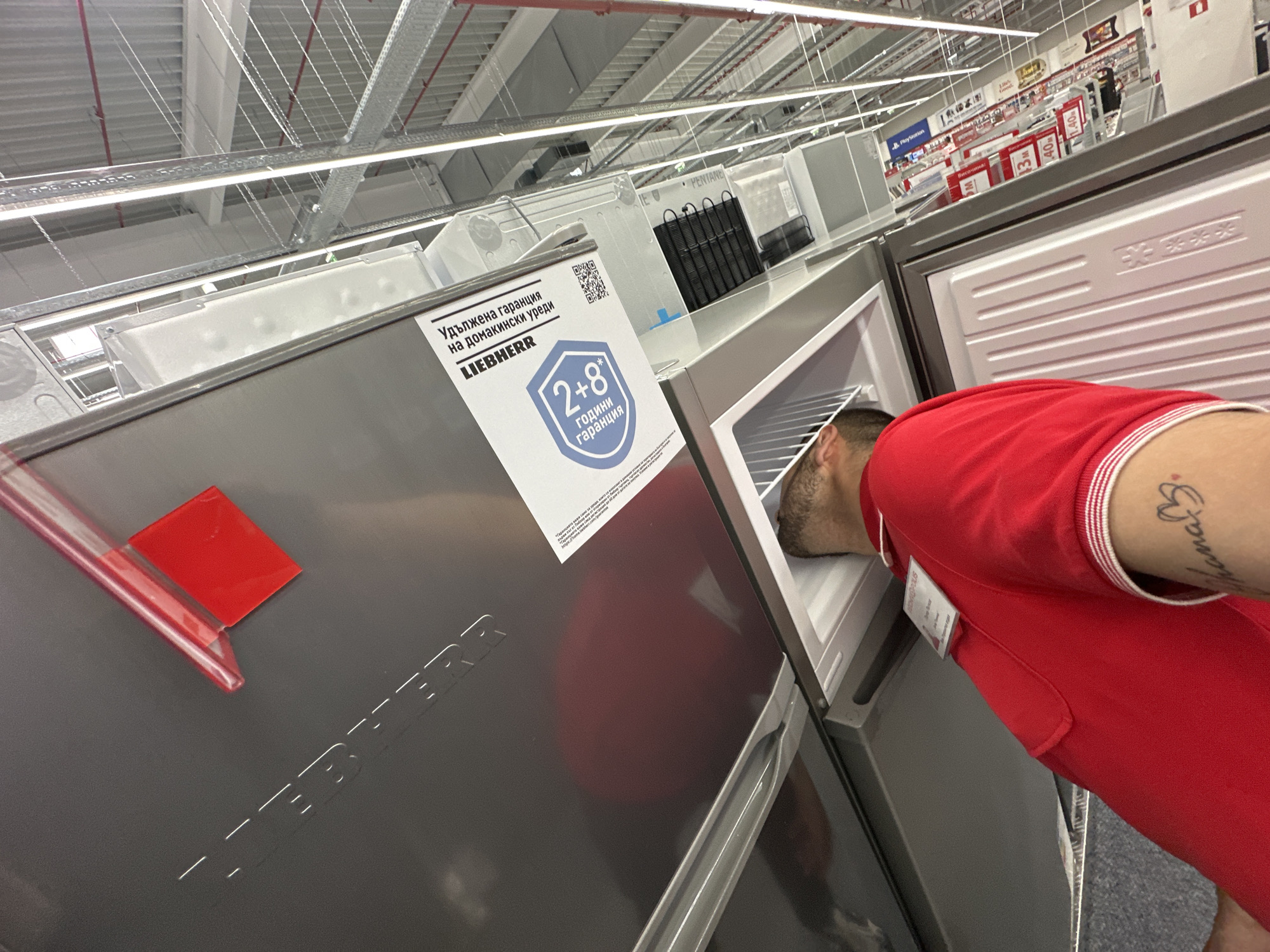 Here's a detailed description of the image:
**Scene:** The photo appears to be taken inside a large appliance store, likely in Lovech, Bulgaria. The environment is characterized by high ceilings with visible industrial-style wiring and support structures, typical of a retail space. A variety of refrigerators and freezers are displayed in rows.
**Objects:**
* **Refrigerators/Freezers:** Several refrigerators/freezers are prominently featured. One is an open freezer with a man's head inside. The brand "Liebherr" is visible on one of the units, along with a promotional sign in Bulgarian advertising a "2+8 years guarantee." There are stickers with the word PENTANE written on them. There are product displays with the word "PlayStation" written on them.
**People:**
* **Man:** A man is looking inside the freezer. He is wearing a red collared shirt and has a tattoo on his arm.
**Actions/Interactions:**
* The man is examining the inside of a freezer, possibly comparing models or checking features before making a purchase.