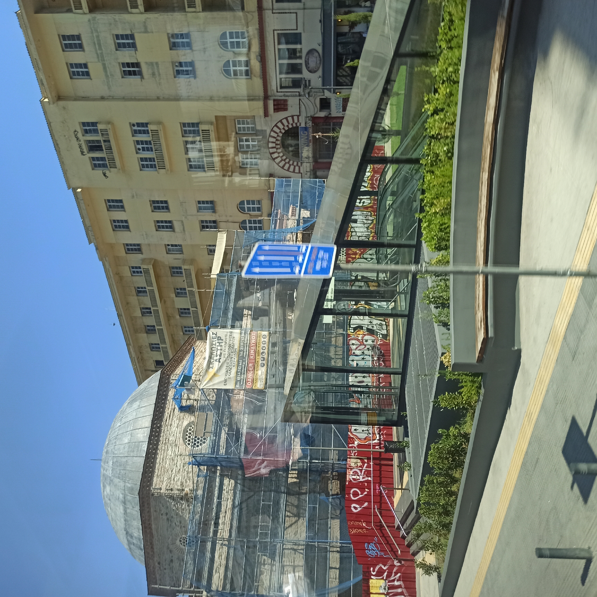 The image depicts a street scene in Thessaloniki, Greece. In the foreground, a wide sidewalk with tactile paving runs horizontally. A wooden bench is situated on a raised planter bed filled with greenery. Behind the bench, a modern glass-enclosed structure with escalators is visible. A prominent blue traffic sign with white arrows pointing upwards is mounted on a pole in front of the glass structure. The middle ground features a section of a building with graffiti-covered red metal walls and stairs leading up to the glass structure. To the left, a domed building, appearing to be a mosque, is partially covered by scaffolding and blue netting, suggesting ongoing construction or renovation. A banner with Greek text is attached to the scaffolding. Behind the mosque, a multi-story yellow building with balconies and numerous windows dominates the background. The sky is clear and blue.