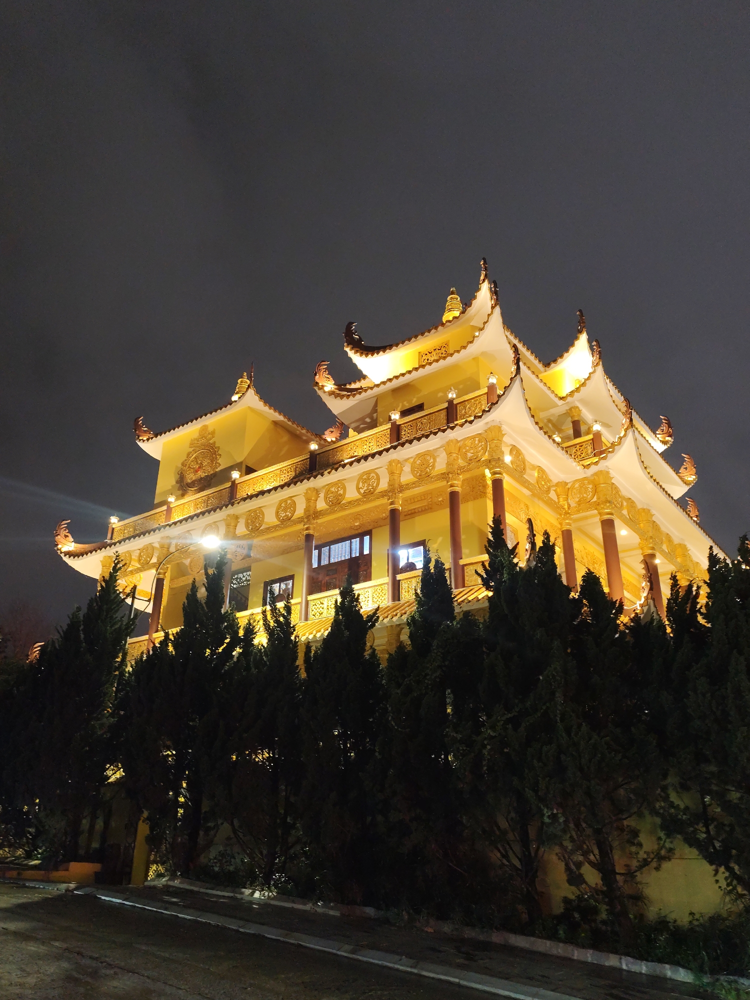 This night-time photograph showcases a magnificent, multi-tiered building, likely a temple or pagoda, brightly illuminated against a dark, overcast sky. The prominent structure features traditional East Asian architecture, characterized by its vibrant golden-yellow facades, elaborate golden ornamentation, and multiple curved, upturned roofs with decorative finials. Warm, artificial lights strategically highlight the intricate details on its balconies, supporting columns (some appearing reddish-brown), and decorative patterns across several visible levels.
In the foreground, a dense row of tall, dark green evergreen trees partially obscures the lower section of the building, forming a natural screen. A dimly lit paved path or road, with a distinct curb, is visible at the bottom left, suggesting an approach to the structure. The dark, cloudy sky indicates it is night. There are no people visible, and no specific activity is taking place. The grand architectural style and serene atmosphere are consistent with a religious or cultural site in Da Lat, Vietnam. No visible text is present in the image.