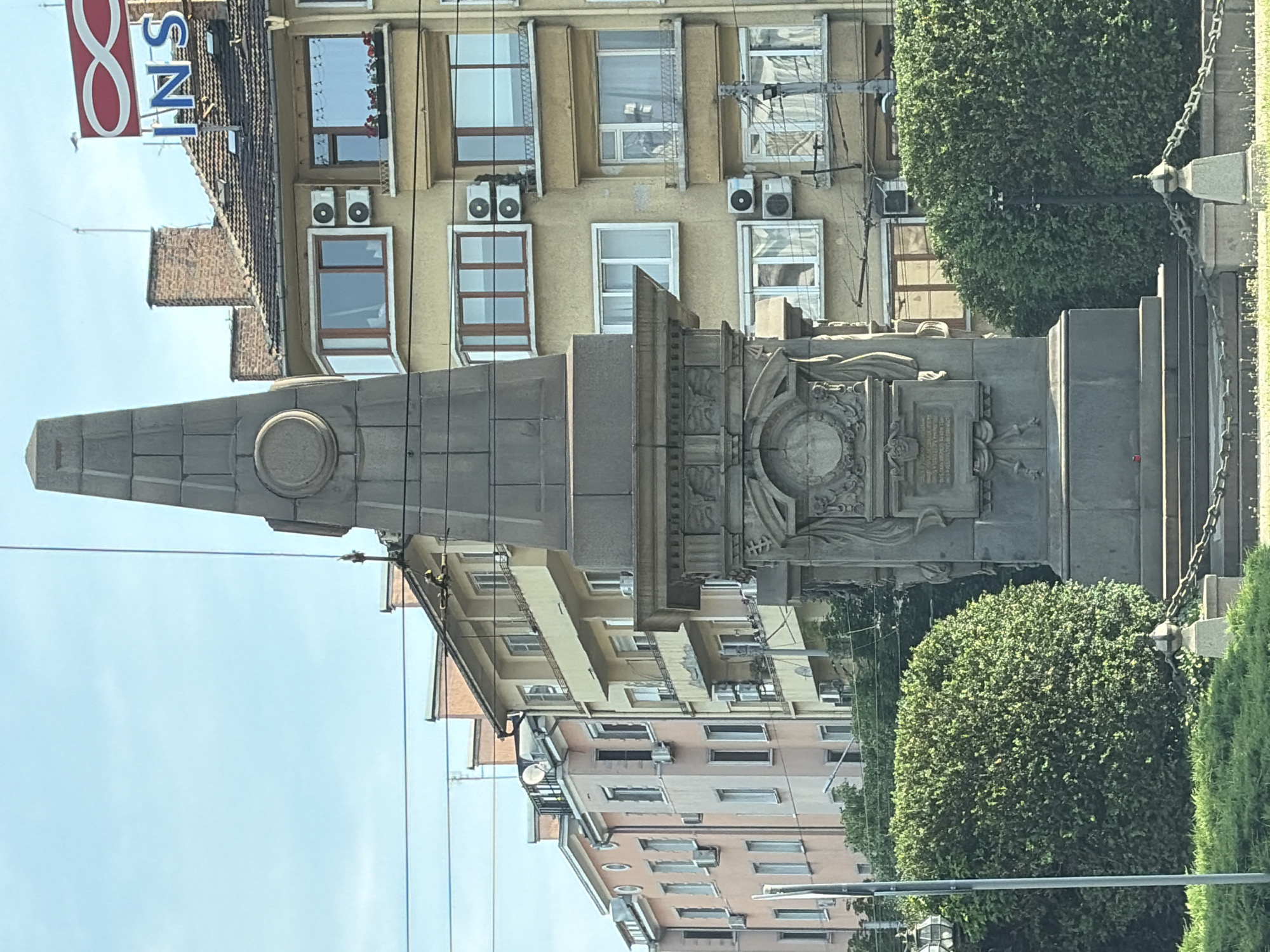 This image depicts an urban street scene in Sofia, Bulgaria, centered on the Monument to Vasil Levski. The monument, a grey stone obelisk, occupies the central foreground. Its base is intricately carved with relief sculptures, including a lower circular medallion displaying a profile bust and a rectangular plaque below it. A simpler circular medallion is visible higher on the obelisk shaft. The immediate area around the monument's base is a patch of green grass and dense shrubs, demarcated by a low chain barrier supported by short stone posts.
In the background, a row of multi-story buildings extends across the frame. To the right of the monument, a light yellow building with a dark tiled roof features a prominent rooftop sign displaying a red infinity symbol alongside the blue letters "INS." This building has multiple windows, some bay-style, some with small balconies and flower boxes, and numerous exterior-mounted white air conditioning units. To the left, a sequence of residential buildings with facades in shades of pink, cream, and light yellow are visible, also exhibiting numerous windows and several air conditioning units. Overhead electrical or communication wires crisscross the upper portion of the scene against a clear, light blue sky. No individuals are visible.