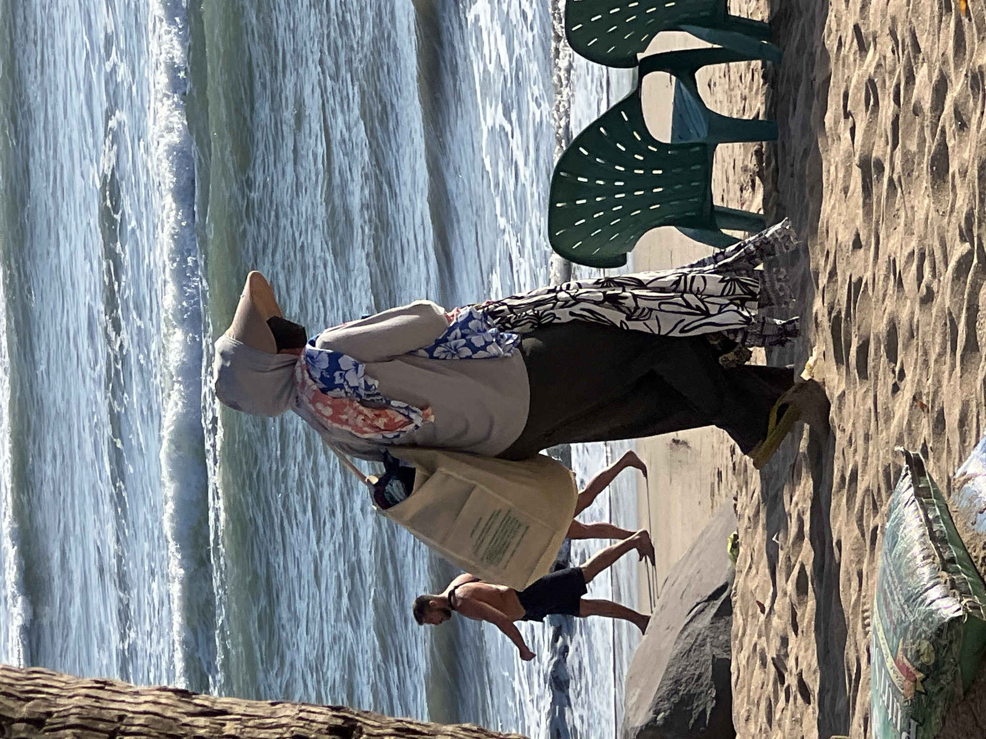 The image depicts a beach scene in Kuta, Indonesia. In the foreground, a person wearing a hooded garment, a wide-brimmed hat, and dark pants walks towards the right side of the frame. A large tote bag is slung over their shoulder, and colorful fabric, possibly a scarf or sarong, is draped over the bag and their shoulder. To the left of this person, a man wearing dark shorts is walking towards the left, with the ocean waves and shoreline visible behind him. Two green plastic chairs are positioned on the sand in the mid-ground, facing the ocean. The sand in the foreground is textured with footprints and there is a large rock formation on the left side of the beach. The ocean in the background exhibits moderate waves with white foam.