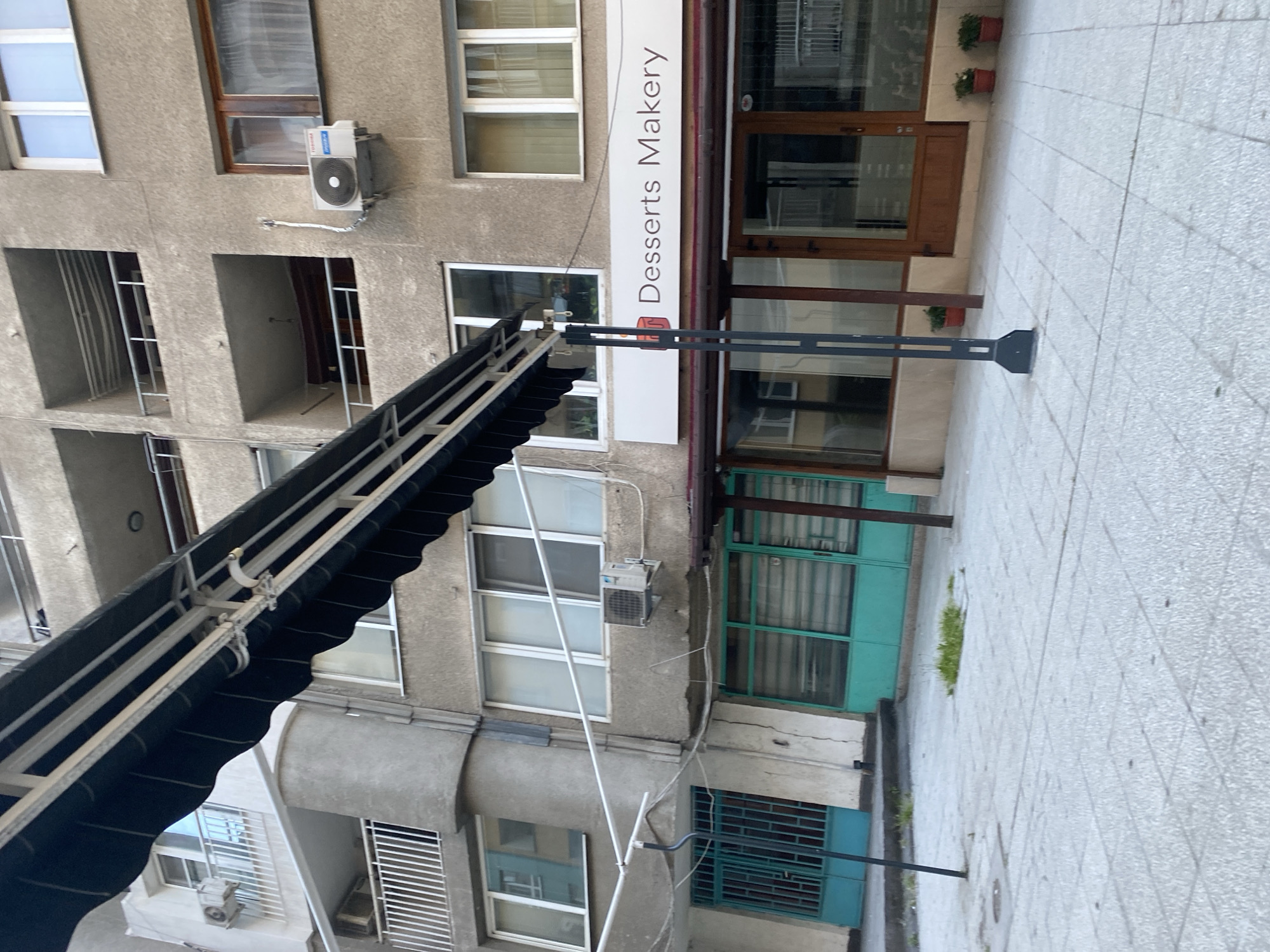 The image shows a view of a building in Sofia, Bulgaria. A sign on the building reads "Desserts Makery". The building has multiple windows of varying sizes and styles. Some windows have bars, while others do not. There are air conditioning units mounted on the exterior walls. A retractable awning extends outward from the building. The surrounding street is paved with grey stone tiles.