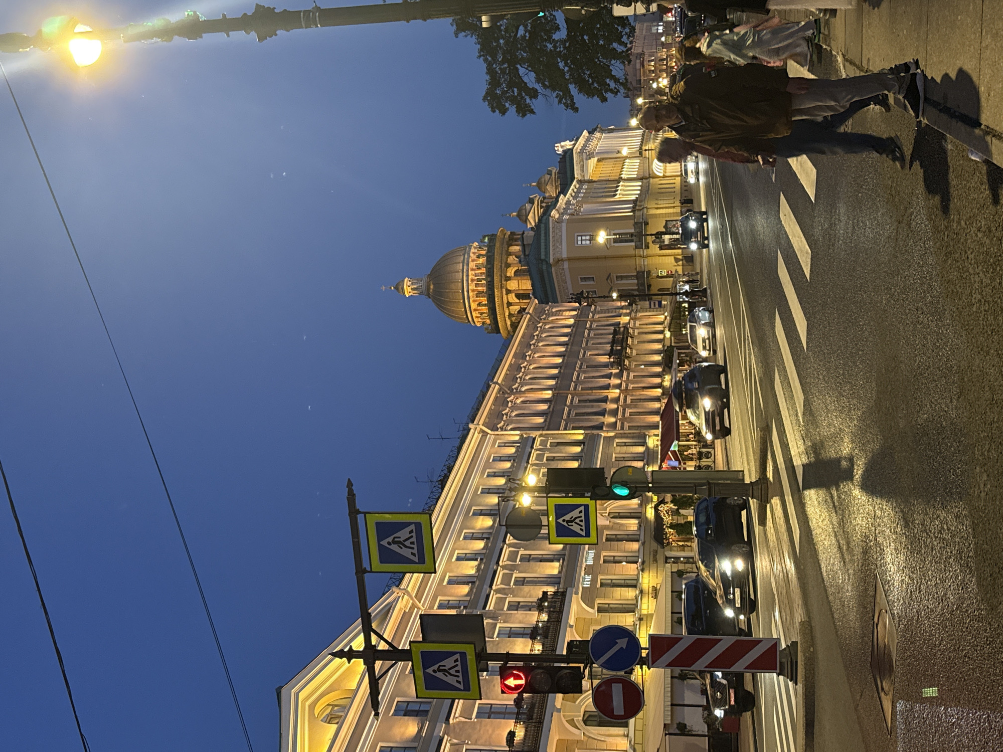 This is a nighttime exterior shot of a street scene in Saint Petersburg, Russia, with the distinctive dome of St. Isaac's Cathedral visible in the background. The image captures a wide street with multiple lanes of traffic and pedestrian crosswalks. Buildings with classical architecture, illuminated with warm yellow lights, line one side of the street. These buildings appear to be a mix of commercial and residential establishments, with some signage visible, including "HOTEL".
Several cars are on the road, some with headlights on, suggesting ongoing traffic. Pedestrian crossing signals are displayed, with a red light indicating vehicles should stop. Traffic lights and street signs, including pedestrian crossing signs and a "no entry" sign, are prominent. On the sidewalk, several individuals are present, some walking and others standing. Two men are in the foreground, appearing to be conversing or observing the scene. The sky is a deep twilight blue. The overall atmosphere is urban and active, characteristic of a city at dusk or early evening.