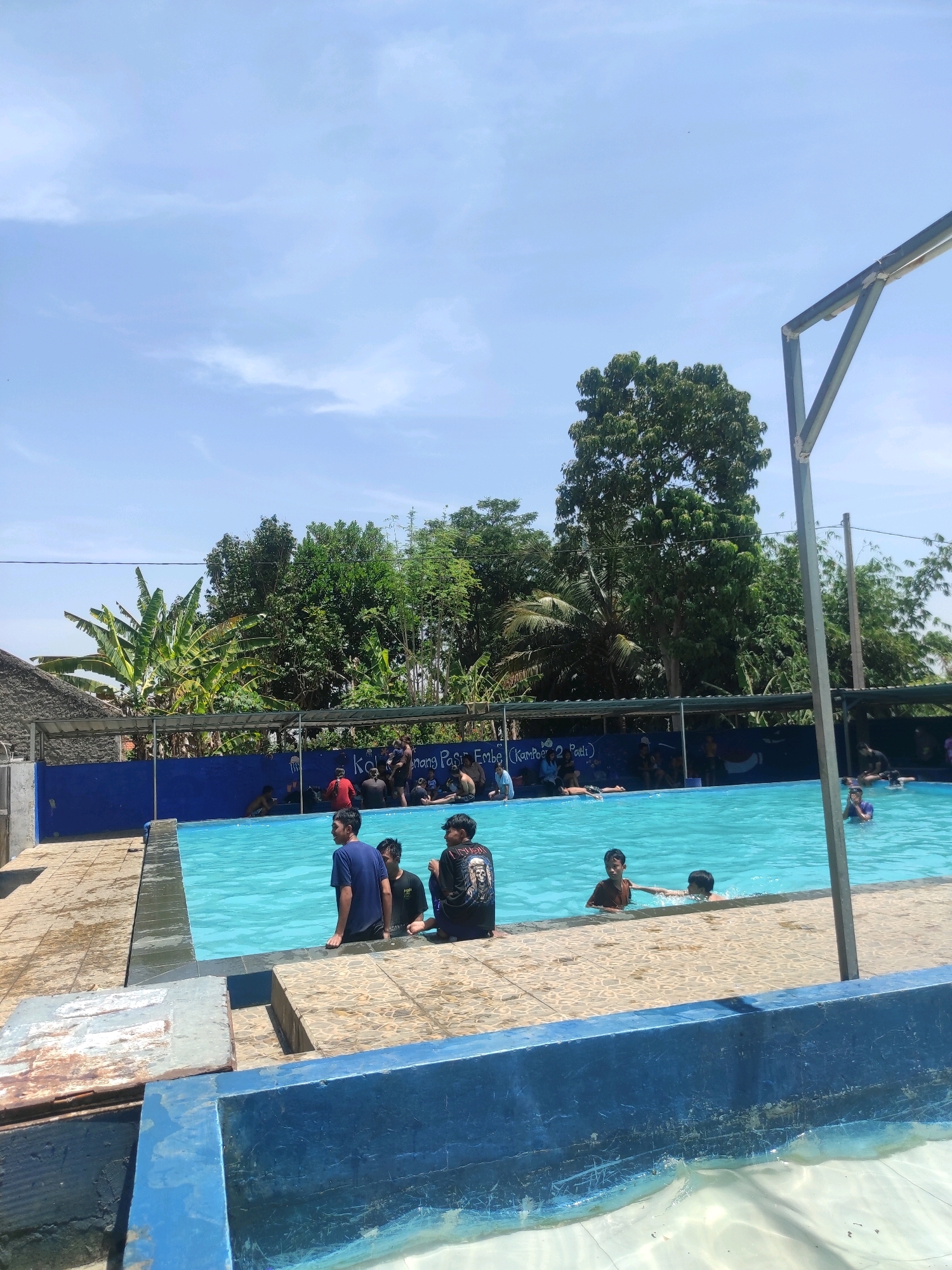 A photograph taken from a slightly elevated angle captures an outdoor swimming pool on a sunny day in Ngamprah, Indonesia. The pool is filled with clear, turquoise water, and several people are visible enjoying it. In the foreground, three young males are standing at the edge of the pool, two facing towards the camera and one with his back to it. To the left, a tiled area surrounds the pool, with some discarded metal sheeting and a weathered blue structure. Further back, a blue-painted wall with a dark blue border runs along the side of the pool, adorned with graphics and text, including "Keren Bang Pas Ecko (Kampos 2 Pati)".
Beyond the pool, lush green trees and vegetation form a dense backdrop. A rustic, thatched-roof structure is visible on the left. The sky above is a bright, clear blue with a few wispy white clouds. The overall scene depicts a casual, recreational setting at a local swimming pool.