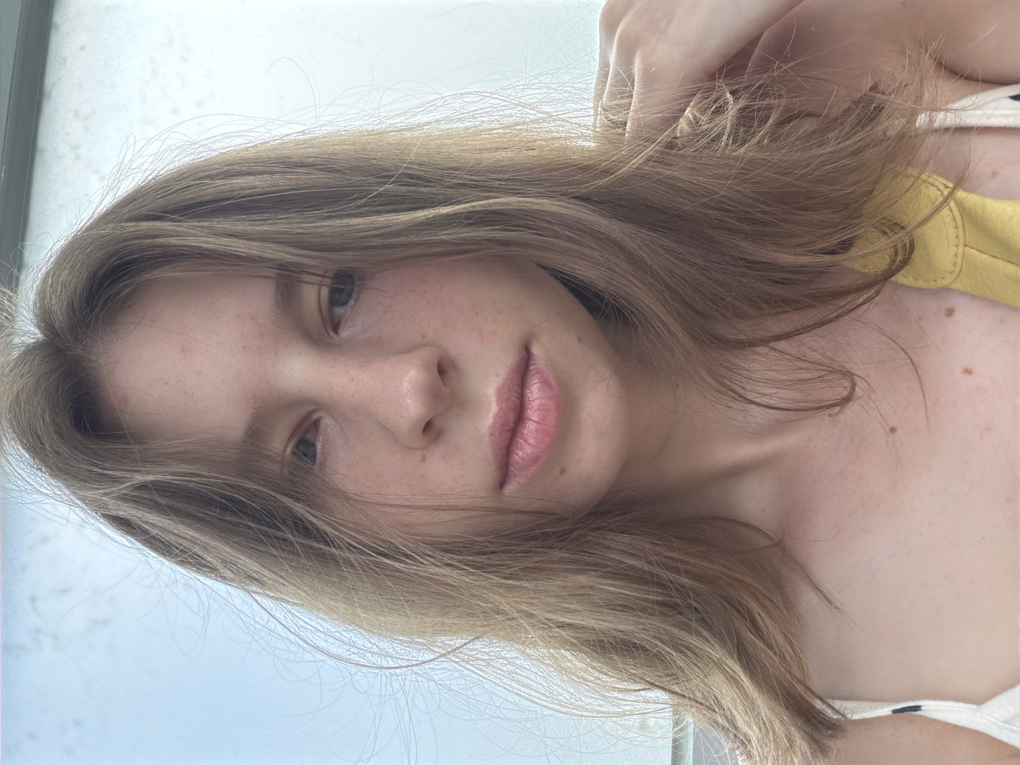 The image shows a close-up, frontal view of a young woman with light brown, shoulder-length hair, fair skin with scattered freckles, and light blue eyes. She is looking directly at the camera. Her lips are slightly parted and appear to be glossy. She is wearing a white top with thin straps, and a hint of a yellow garment is visible. Her left shoulder and a portion of her upper chest are also visible. The background is blurred and appears to be an outdoor setting with a light sky. There is no discernible city of Dongducheon-si, South Korea.