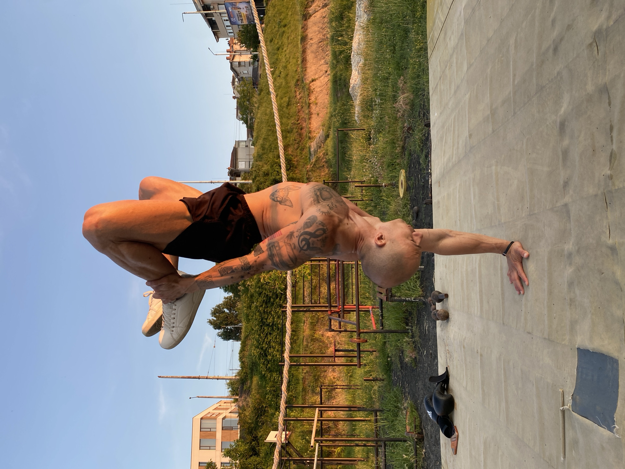 In Pleven, Bulgaria, an athletic individual is performing a handstand on a raised platform during sunset. The person, with numerous tattoos, is wearing black shorts and white sneakers. Their body is positioned vertically, with one hand supporting their weight and the other holding their leg. The background features a grassy hill with an outdoor gym structure, some buildings, and a clear blue sky. On the ground near the platform are boxing gloves, a smartphone, and weights. The warm light of the setting sun casts a golden hue over the scene.