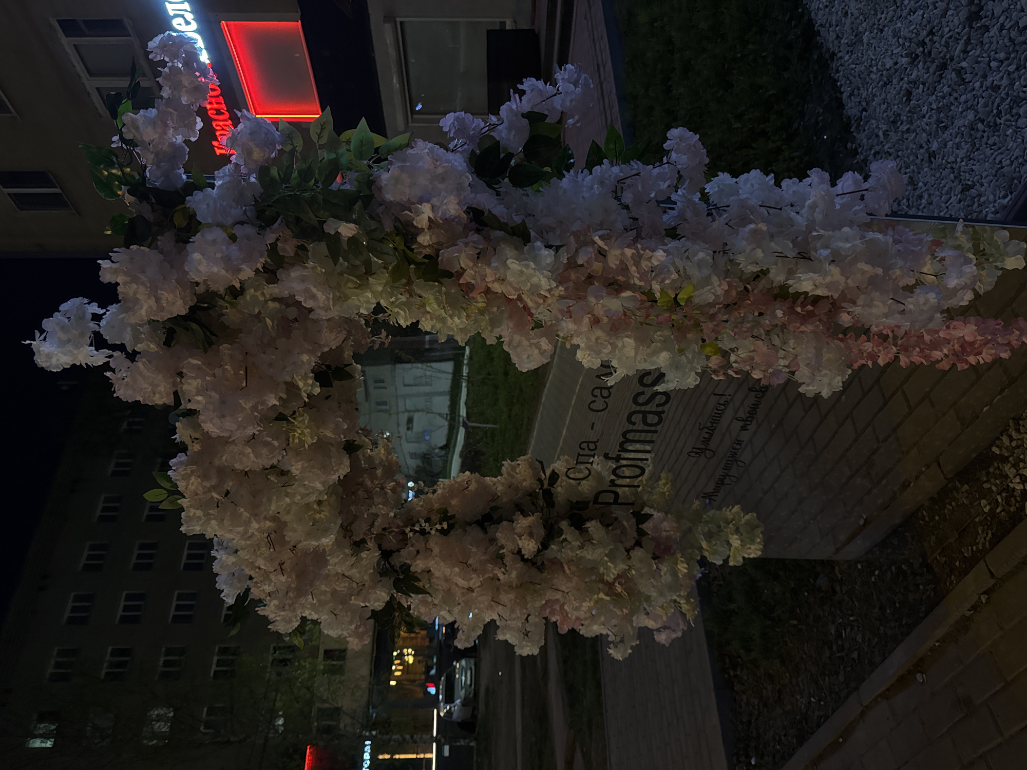 A nighttime outdoor shot captures a floral display adorning a sign that reads "Спа - салон Profmass", situated in Izhevsk, Russia. The sign is predominantly white, its text in Cyrillic script, with "Profmass" appearing to be the name of the establishment.
The main focus is a decorative arrangement of white and light pink flowers, possibly artificial, which surrounds the sign. Some green leaves are also visible within the floral arrangement. The flower wreath is shaped like a frame.
Buildings are visible in the background, including what appears to be an apartment building with multiple windows lit up. The streetlights and neon signage add to the urban ambiance of the scene. Sidewalk is also visible. A red sign is seen on one of the buildings. The setting appears to be a city street or a sidewalk in an urban area.
