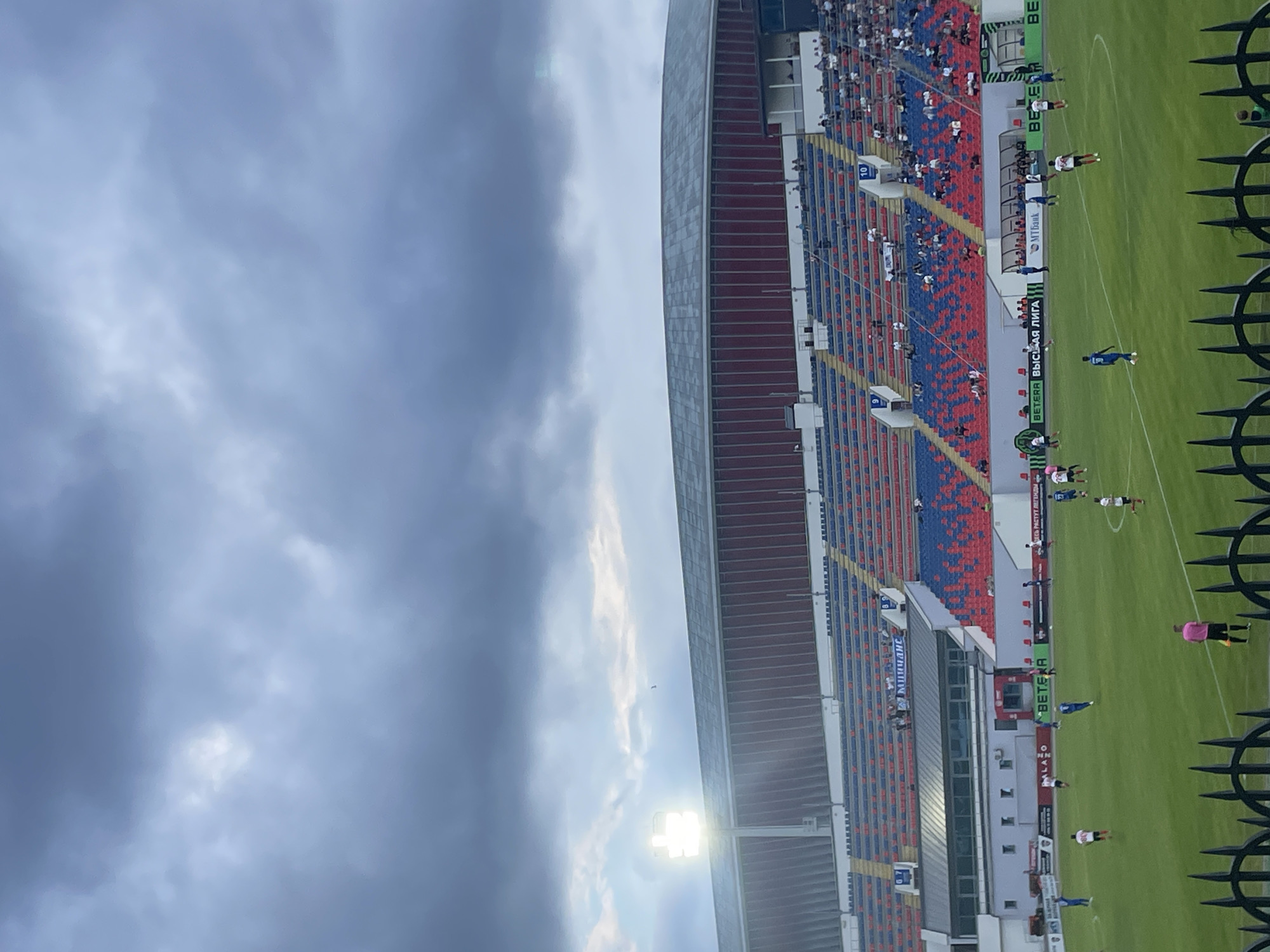 This is a vertical, eye-level shot of a football stadium with an overcast sky and a bright stadium light shining. The stadium seating is visible, with rows of red and blue seats. Some spectators are seated in the stands, appearing as small figures from this distance. On the green football field, several players in white and blue uniforms are engaged in a game. The scene suggests an outdoor sporting event.
Notable details include the prominent stadium light, which is illuminated, indicating it might be evening or that artificial lighting is needed due to the cloudy conditions. The sky is largely filled with gray clouds, suggesting a potentially overcast or stormy day. The presence of advertising boards around the field, some with visible text, points to a professional or organized sporting venue.
Visible text includes "BETEPA" and "ВЫСШАЯ ЛИГА" (Vysshaya Liga, meaning "Premier League" in Russian) on advertising boards around the field. There is also text that appears to read "MTБaнк" (MTBank). The overall atmosphere is one of a sports match in progress, under a dramatic sky. The image captures the scale of the stadium and the activity on the field, contrasting the man-made structure with the natural elements of the sky.