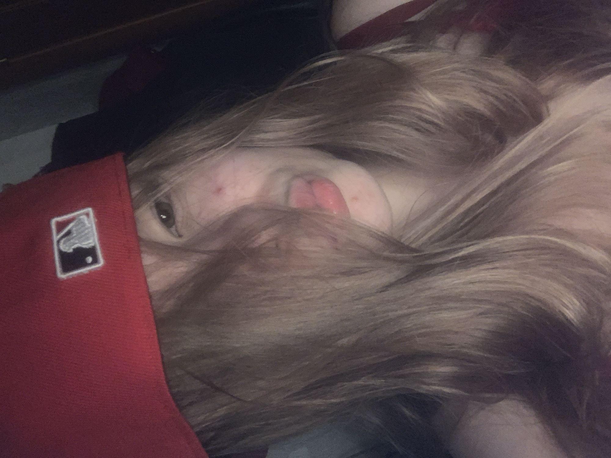 A person with long, light brown hair is lying down wearing a red baseball cap with the MLB logo. The person's face is partially obscured by their hair and the cap. Only one eye is visible. The person's mouth is slightly open, revealing their tongue. The background is dark and indistinct. The image does not contain any information suggesting the location is in the city of Saint Petersburg, Russia.