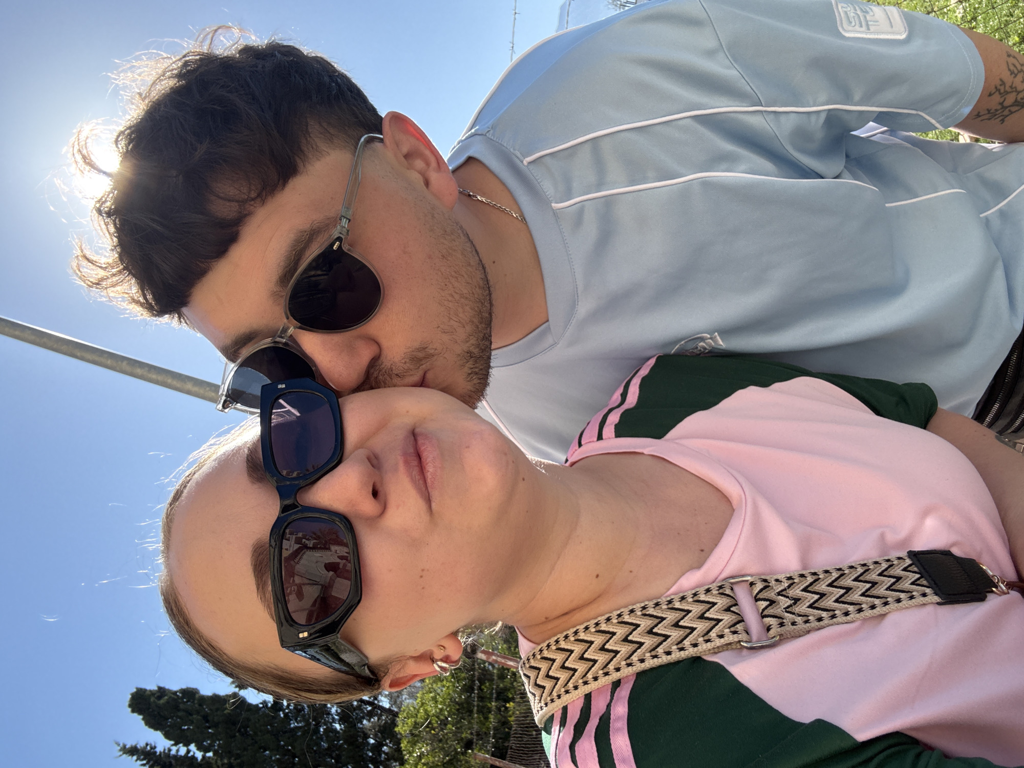 A selfie depicts a man kissing a woman outdoors. The man has dark hair and a beard, and is wearing sunglasses and a light blue t-shirt. He is kissing the woman on the forehead. The woman has light hair and is also wearing sunglasses. She is wearing a pink and green shirt with a patterned strap. The background features a blue sky, the sun, trees, and architectural elements. Based on available information, the photo was likely taken outdoors in Bar, Montenegro.