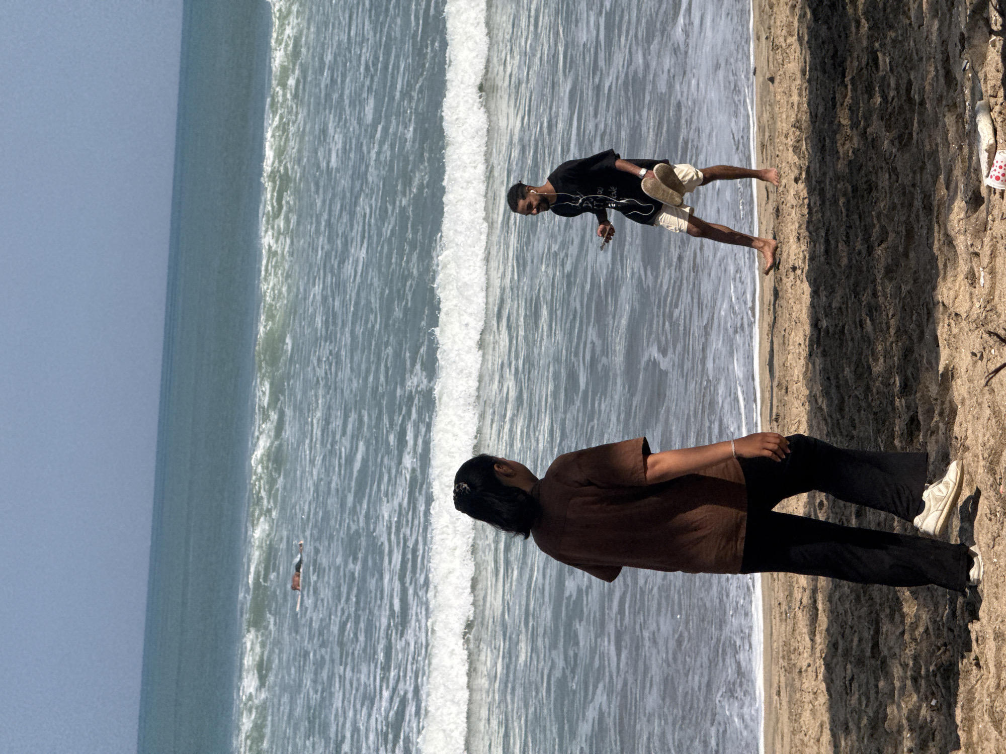Two individuals are on a sandy beach in Kuta, Indonesia, with the ocean and waves in the background. In the foreground, a person with dark hair tied in a bun, wearing a brown t-shirt and dark pants, stands facing away from the camera, looking towards the ocean. To their right and slightly further back, a person with short dark hair and a beard, wearing a black t-shirt and light-colored shorts, is holding a smartphone and earbuds in one hand and a pair of flip-flops in the other. They are looking to their left, with a slight smile. In the middle distance, a person is lying on a surfboard in the ocean. The sky is clear and blue. The sand is a light brown color, and there are some shadows cast on it. The water is a deep blue-green with white-capped waves.
