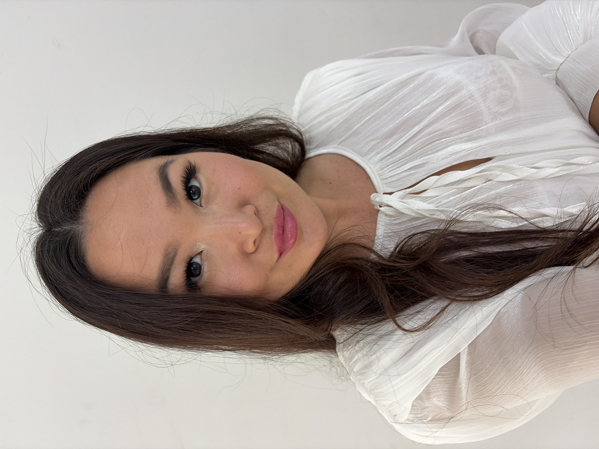 A woman with long, dark hair and fair skin poses for a portrait against a plain, off-white background. She is wearing a white, semi-sheer blouse with a ruffled neckline and braided ties. Her makeup includes dark eyeliner, mascara, false eyelashes, blush, and glossy pink lipstick. The lighting is even, and the background suggests an indoor setting, possibly a home or studio. There is no indication of the city of Chaikovsky, Russia, in the image.