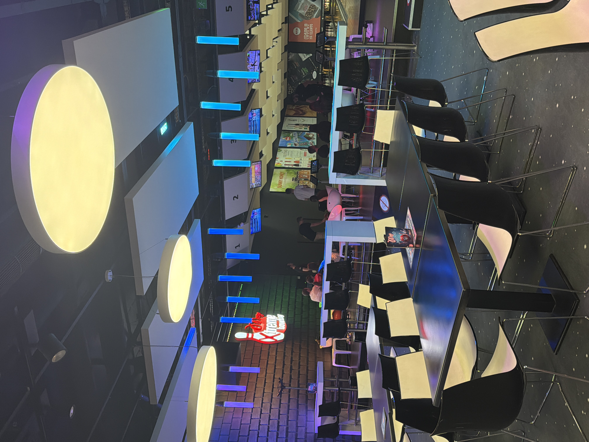 The image depicts the interior of an establishment, possibly a bowling alley or entertainment venue, in Sofia, Bulgaria. It showcases long, dark tables with black and white chairs, likely arranged for dining or socializing. Above, there are circular, illuminated lights and rectangular panels suspended from the ceiling. The backdrop features numbered screens or lanes, and displays of advertisements and products. People can be seen in the background, some standing or sitting. The overall atmosphere appears modern and illuminated, suggesting a recreational space.