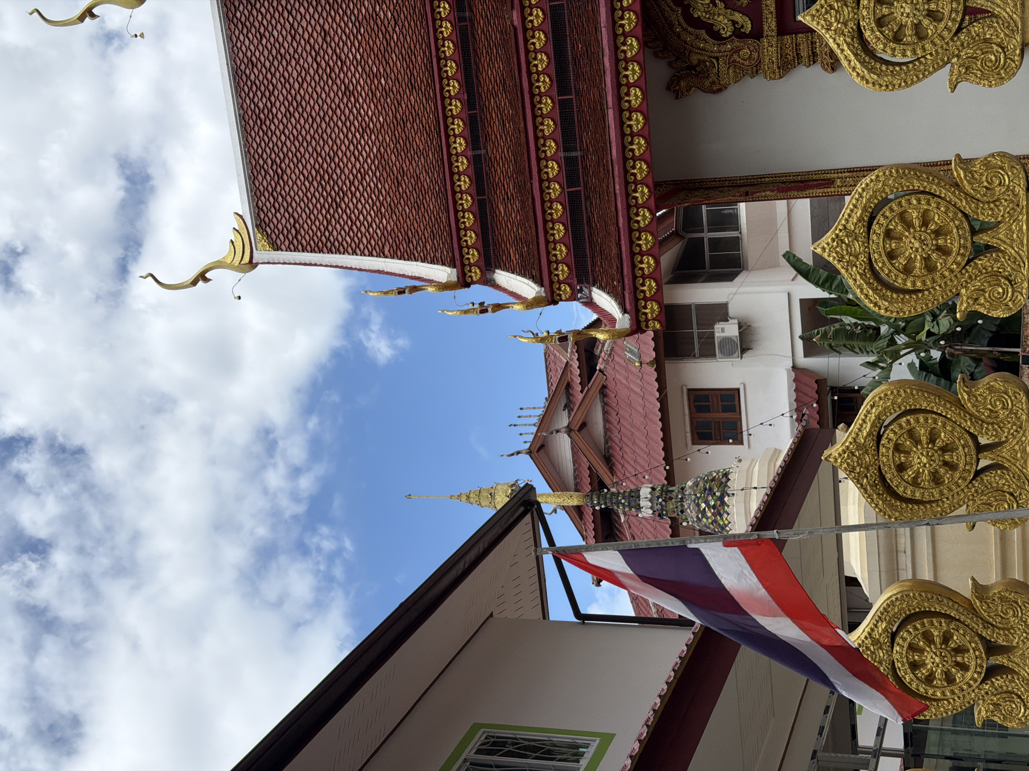 The image captures a view of a temple complex in Chang Phueak, Thailand, on a partly cloudy day. The foreground features a vibrantly colored Thai flag, its red, white, and blue stripes stark against the sky. To the right of the flag, ornate golden decorative elements, resembling stylized lotus flowers with dharma wheels at their centers, are prominently displayed.
Dominating the right side of the frame is the roof of a temple building, characterized by its deep red, tiled surface and intricate golden trim. The roof is adorned with decorative golden finials shaped like mythical creatures. Adjacent to this, and further into the background, are other structures with similar red roofs and golden accents, suggesting a larger temple complex. One building has a visible air conditioning unit mounted on its exterior wall.
Behind these buildings, the sky is a mix of bright blue and white cumulus clouds. In the distance, a tall, multi-tiered golden stupa rises, a common architectural feature in Thai Buddhist temples. The overall impression is one of rich cultural and religious architecture, set against a typical Thai sky. The image is static, with no people or animals visible, and no discernible actions or interactions taking place. The environment is one of clear urban architecture, likely a tourist or religious site.