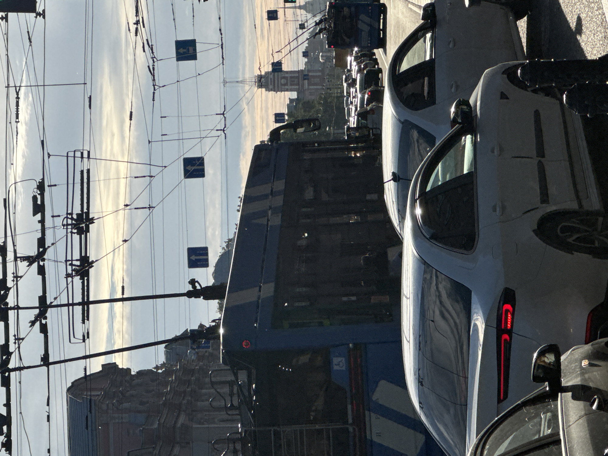 This media file captures a street scene in Saint Petersburg, Russia, likely during the day as indicated by the bright sky and shadows. The image is oriented vertically, suggesting it was taken from a moving vehicle or with a camera held in a portrait orientation.
The primary subjects are vehicles and infrastructure. In the foreground, a white car is prominently featured, its sleek design and modern taillight visible. Behind it, another car is partially visible. A blue bus or tram dominates the middle ground, with a person visible inside through the darkened windows. The bus/tram appears to be in traffic, as are the cars behind it. The background reveals more traffic, including a line of cars that stretch into the distance. Tall buildings, characteristic of Saint Petersburg, rise in the background, including a prominent clock tower. Overhead, a complex network of wires and poles, likely for trams or trolleybuses, crisscrosses the sky.
Two blue traffic signs are visible, each displaying a white arrow pointing left, indicating a mandatory turn or lane direction. The sky is partly cloudy, with bright streaks of sunlight suggesting either early morning or late afternoon. The overall atmosphere is that of a busy urban environment. There is no visible text in the image, aside from what might be on the traffic signs, which are not legible.