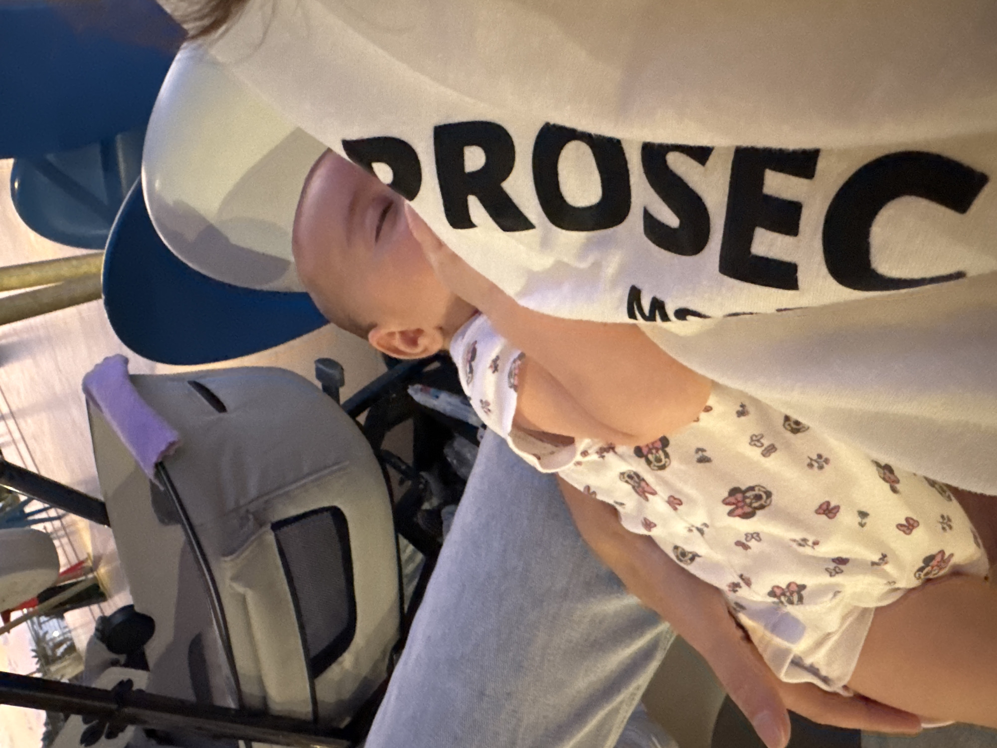The image shows a close-up view of a baby being held, with the back of a person's head and shoulder visible on the right. The baby is wearing a white onesie with a Minnie Mouse print. The person holding the baby is wearing a white shirt with large black text that reads "PROSEC" vertically. The background appears to be indoors, possibly a waiting area or a public space, with glimpses of a stroller and blue chairs. The overall lighting suggests an indoor environment. There is no indication of the city of Isola delle Femmine, Italy.