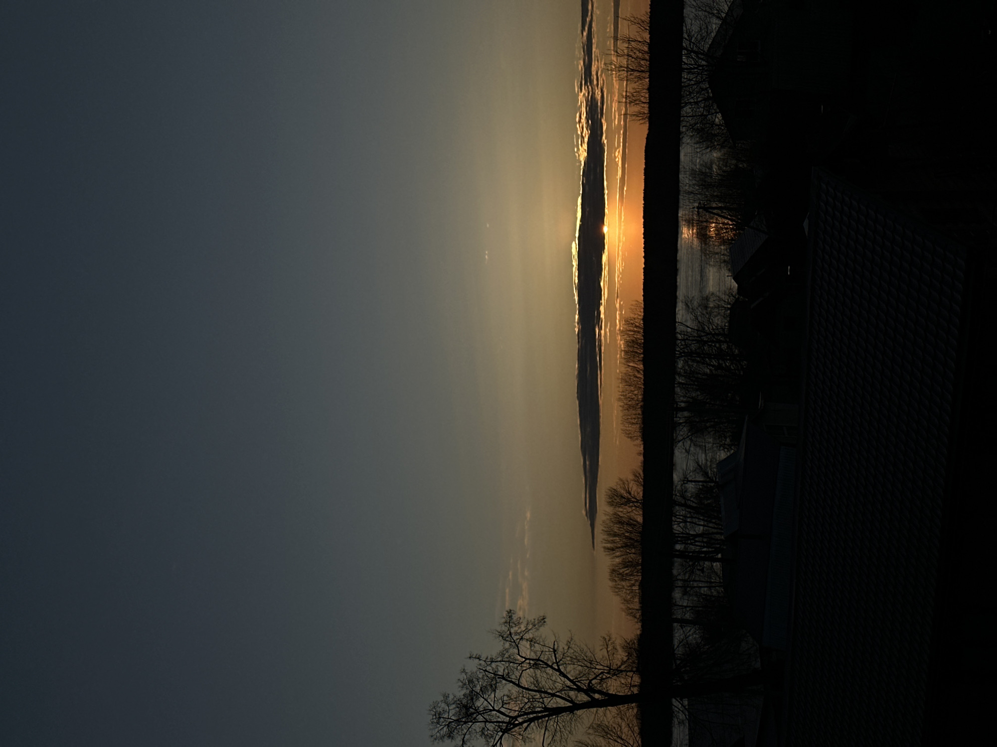 Sunset over the Kama River in Chaikovsky, Russia. Silhouetted trees and a building are visible in the foreground. The sun is setting behind clouds, creating a bright golden light on the water.