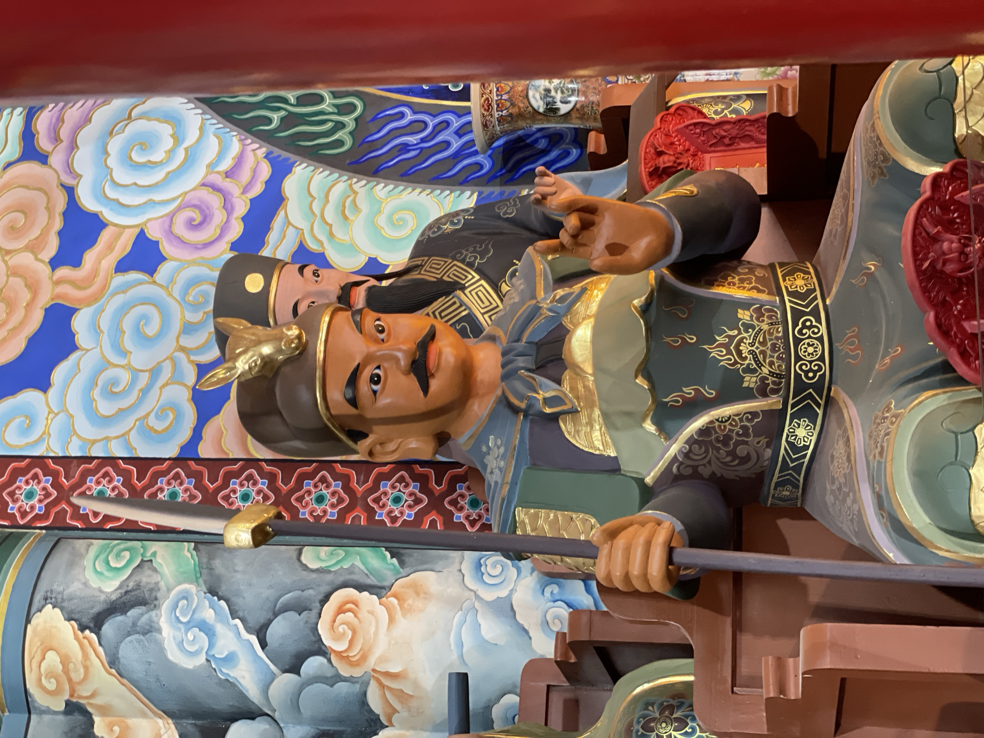 A low-angle, close-up shot depicts a colorful, richly decorated statue of a Chinese warrior figure. The statue is positioned vertically. The warrior has tan skin, a black mustache, and a serious expression. He wears elaborate gold and black armor, and a headdress with a gold animal emblem. The statue is holding a long, dark pole weapon horizontally in both hands.
Behind the warrior, there is a background of painted clouds and other decorative elements in blues, whites, and golds. Another figure is partially visible behind and to the side of the main warrior statue. The second figure has a black hat and a similar tan skin tone.
The statue appears to be located in the city of Lintong District, China.
