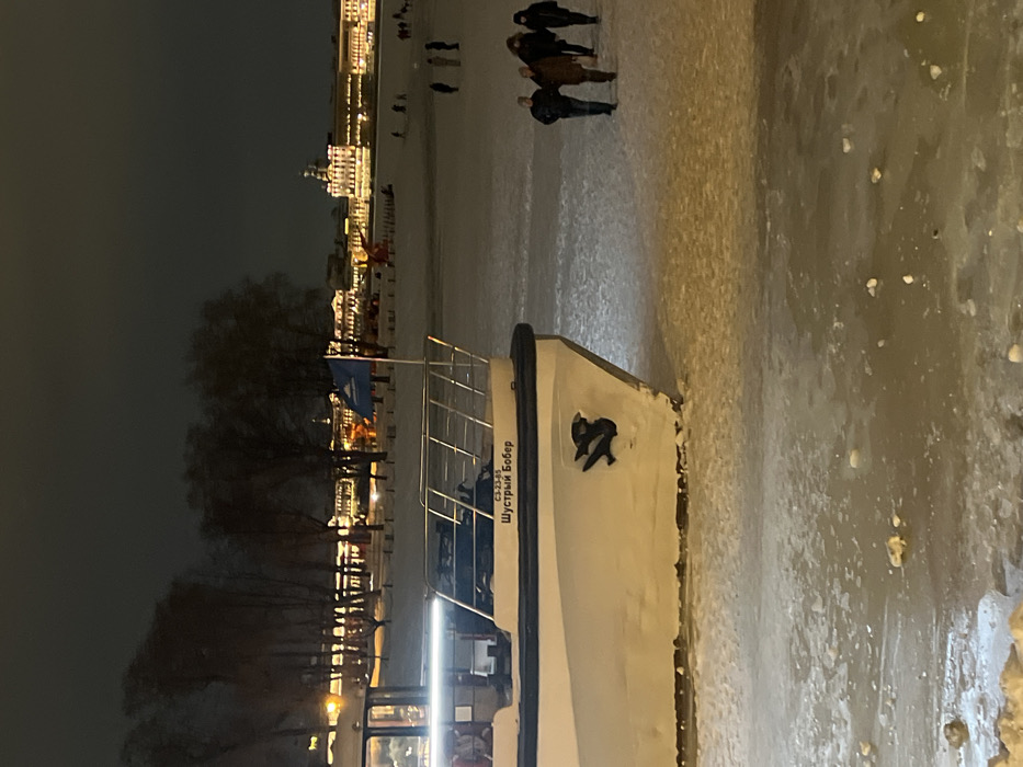 A white boat with black trim, named "Shustryi Bober," sits in a frozen body of water. The anchor is visible on the boat's bow. People stroll on the frozen surface near the riverbank. In the background, illuminated buildings line the shore, adding light to the dark night sky. The scene exudes a chilly, wintry atmosphere.