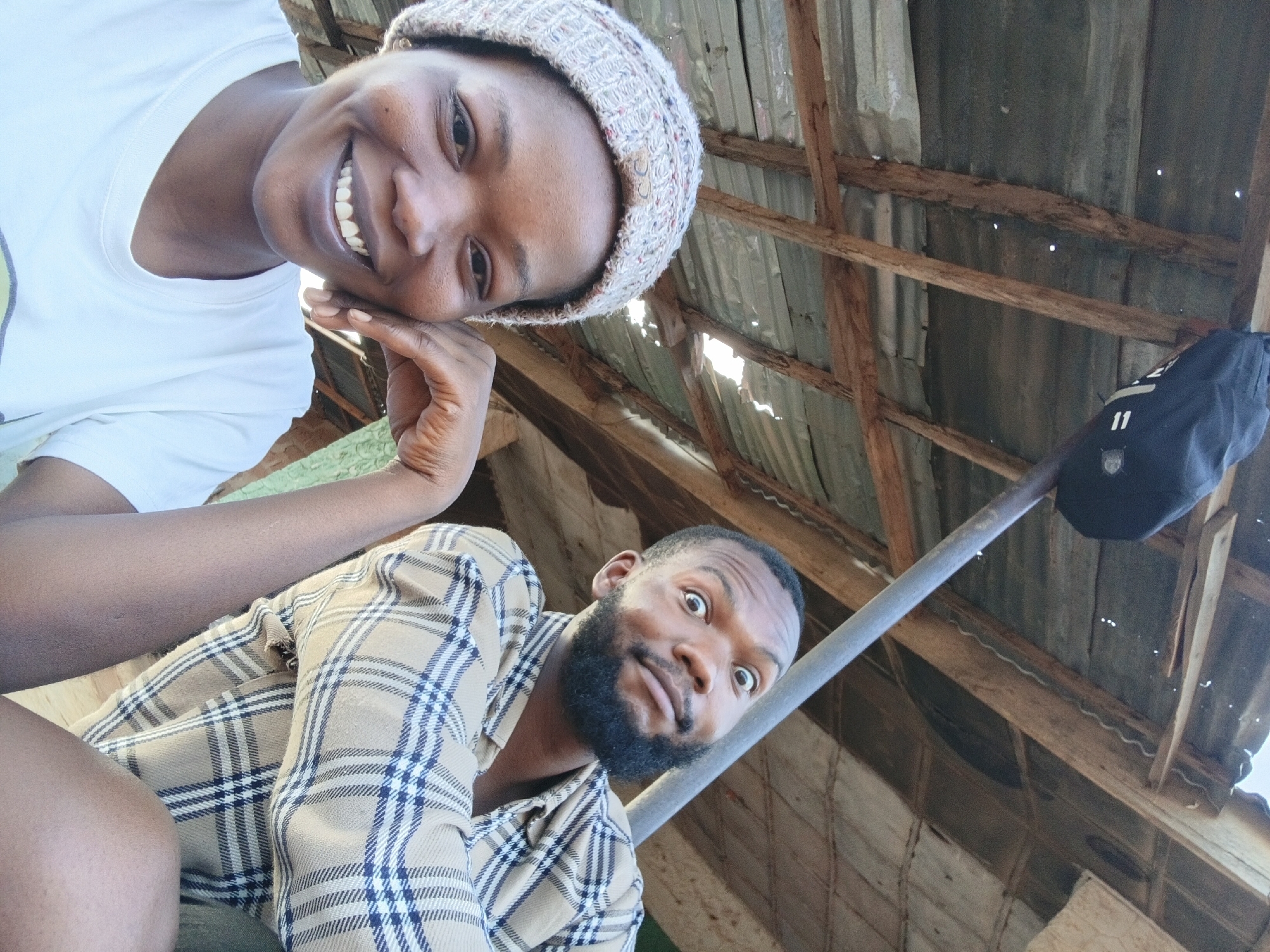 Two individuals are captured in a selfie-style photograph, appearing to be in a partially enclosed structure. The person at the top is a dark-skinned woman wearing a white t-shirt and a knit cap. She is smiling and her right hand is resting against her face. Below and to the right of her is a dark-skinned man with a beard, wearing a plaid shirt. His expression is exaggerated, with wide eyes and raised eyebrows. A metal pole stretches diagonally across the frame, partially obscuring the background. Above and behind the subjects, a roof structure is visible, consisting of corrugated metal sheets and wooden beams. A black bag is attached to the metal pole. The scene appears to be in an outdoor or semi-outdoor setting with natural lighting. The location of the picture is in Jos, Nigeria.