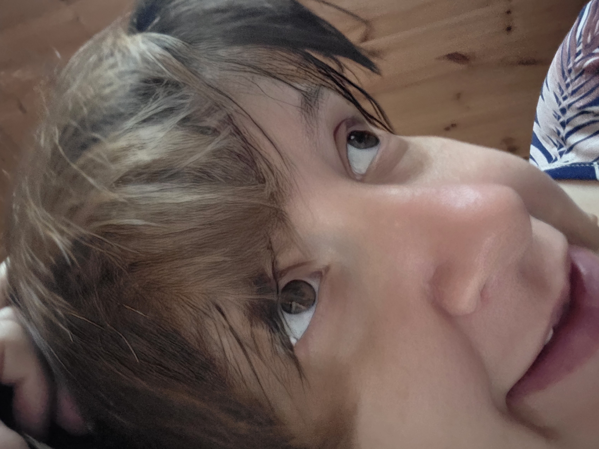 This is a close-up, eye-level, indoor photograph taken in Dunnae-myeon, South Korea. The image features a person's face, with a focus on their eyes and mouth. The person has light skin and short, layered brown and blonde hair. Their right eye, which is light brown, is looking slightly upwards and to the right. Their left eye is also visible and appears to be a similar shade of brown. The person's mouth is open, revealing their teeth and tongue, and they have a slight smile. The background is a blurred wooden surface, suggesting an indoor environment. The lighting is soft and natural, highlighting the person's features.