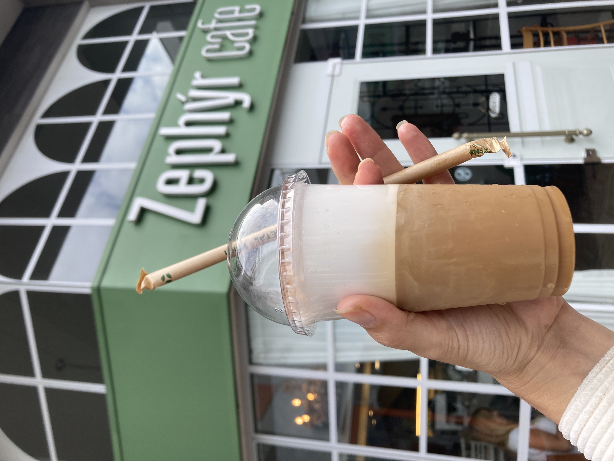 A hand holds a plastic cup of iced coffee in front of a building. The coffee cup is clear with a domed lid. A tan paper straw sticks out of the cup. The drink appears to be layered, with a creamy white layer on top and a tan coffee layer below. The hand holding the cup is pale with neutral nail polish and wearing a white textured sleeve.
Behind the hand and coffee, the green facade of a building is visible. The sign reads "Zephyr cafe" in silver letters. The building has white-framed windows with arched tops, and a white door with a window can be seen.
The location is Florianópolis, Brazil, as evidenced by the "Zephyr cafe" sign, a known business in the area.