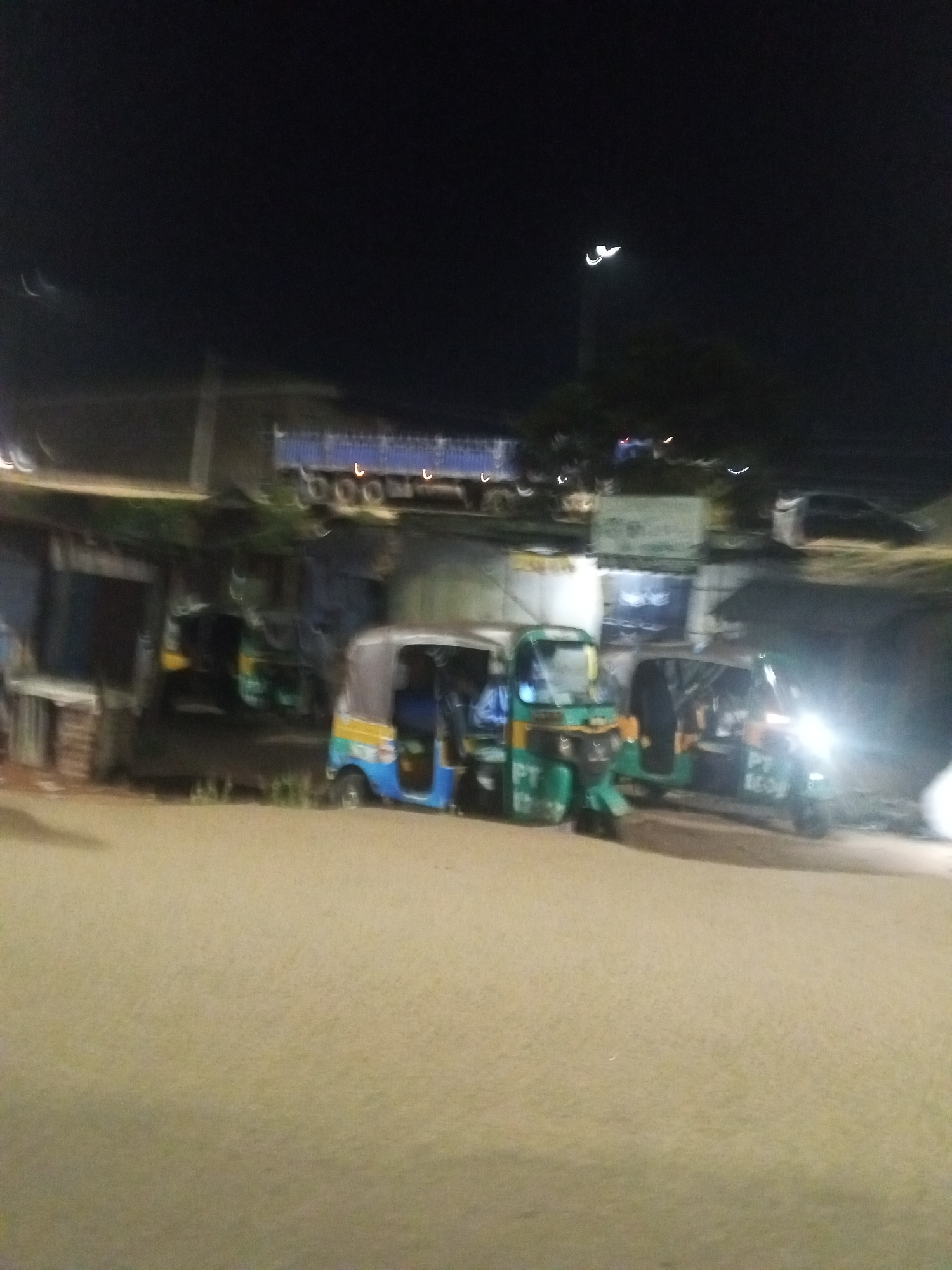 The image shows a street scene at night in Gwong, Nigeria. Three auto rickshaws (tuk-tuks) are parked on the side of a paved road. The one in the center is blue and yellow, while the other two are green. The lights of one of the rickshaws are on. Behind the rickshaws are buildings or shops, and a truck is parked higher up on a platform. The sky is dark. A streetlight is visible in the distance. The road surface is illuminated, but the overall scene is blurry.