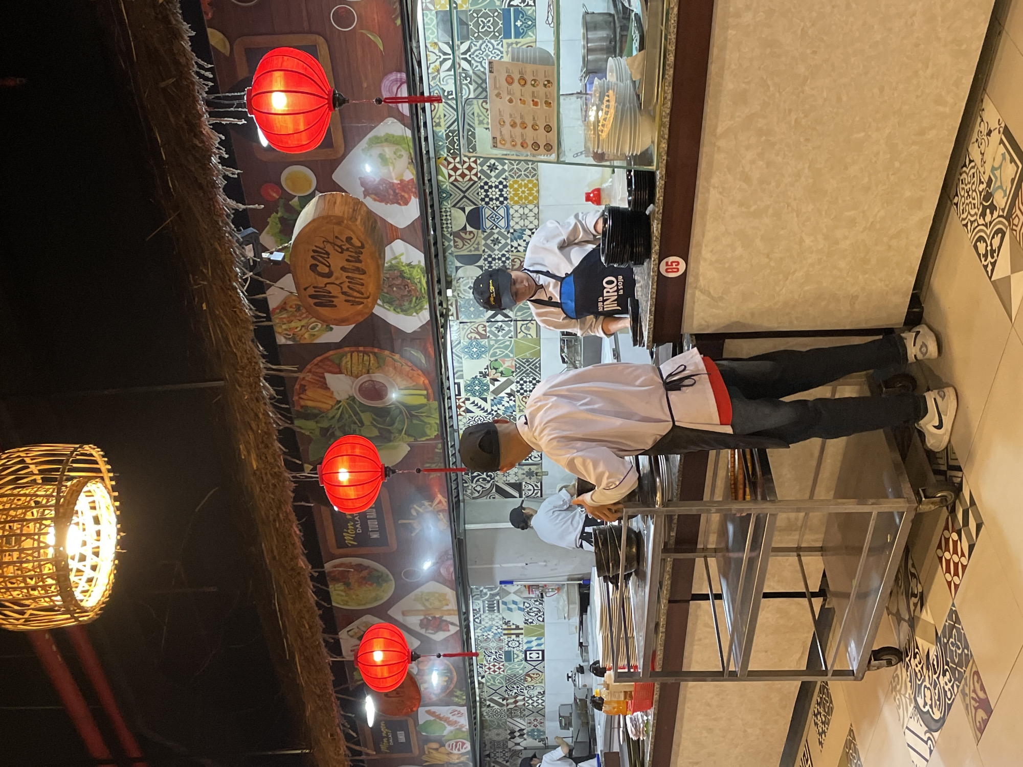 The detailed image depicts an indoor open-plan kitchen or food preparation area within a restaurant in Da Lat, Vietnam. **Location:** The establishment is identified as being in Da Lat, Vietnam, based on a prominent wooden sign on the back wall featuring the text "MÓN NGON ĐÀ LẠT" (Delicious dishes of Da Lat) and "MÌ TƯƠI RAU" (Fresh noodles with vegetables). Another roundish wooden sign in Vietnamese, likely reading "Mì cay Hàn Quốc" (Korean Spicy Noodles), suggests a blend of cuisines. **Environment and Decor:** The ceiling is dark, from which hangs a large, intricately woven bamboo light fixture and several smaller, illuminated red spherical Vietnamese lanterns with tassels. A thatched or straw-like trim extends along the base of the ceiling, above a patterned back wall. This wall features a vibrant mural or large prints depicting various dishes, including what appears to be noodles and salads. Below this, the kitchen backsplash is tiled with a diverse mosaic of patterned squares in shades of blue, green, yellow, and white. The floor is covered in an eclectic mix of geometric and floral patterned tiles. **People:** At least three individuals are visible, all appearing to be kitchen staff. 1. **Foreground Chef:** Positioned on the right with his back mostly to the camera, this male chef wears a white chef's jacket, a dark apron tied at the waist, dark trousers, and a dark baseball cap. He is standing behind a four-tiered stainless steel utility cart, actively handling a dark bowl over a counter. He is wearing white Nike sneakers. 2. **Midground Chef:** Located to the right of the center, this female chef wears a white chef's jacket, a blue apron bearing the "JINRO IS SOJU" logo, and a dark cap. She is focused on tasks at a stainless steel counter, surrounded by stacks of black bowls. 3. **Background Chef:** Partially visible on the far left, another female chef in a white jacket and dark cap is working at a counter further back. **Objects and Equipment:** - The kitchen features stainless steel countertops and various cooking utensils, pots, and pans. - Stacks of clean white plates and black bowls are arranged on the counters. - A stainless steel multi-tiered cart is prominent in the foreground. - A menu board displaying dishes with pictures and text is visible on the right side of the kitchen wall. - A long service counter runs horizontally across the mid-ground, faced with