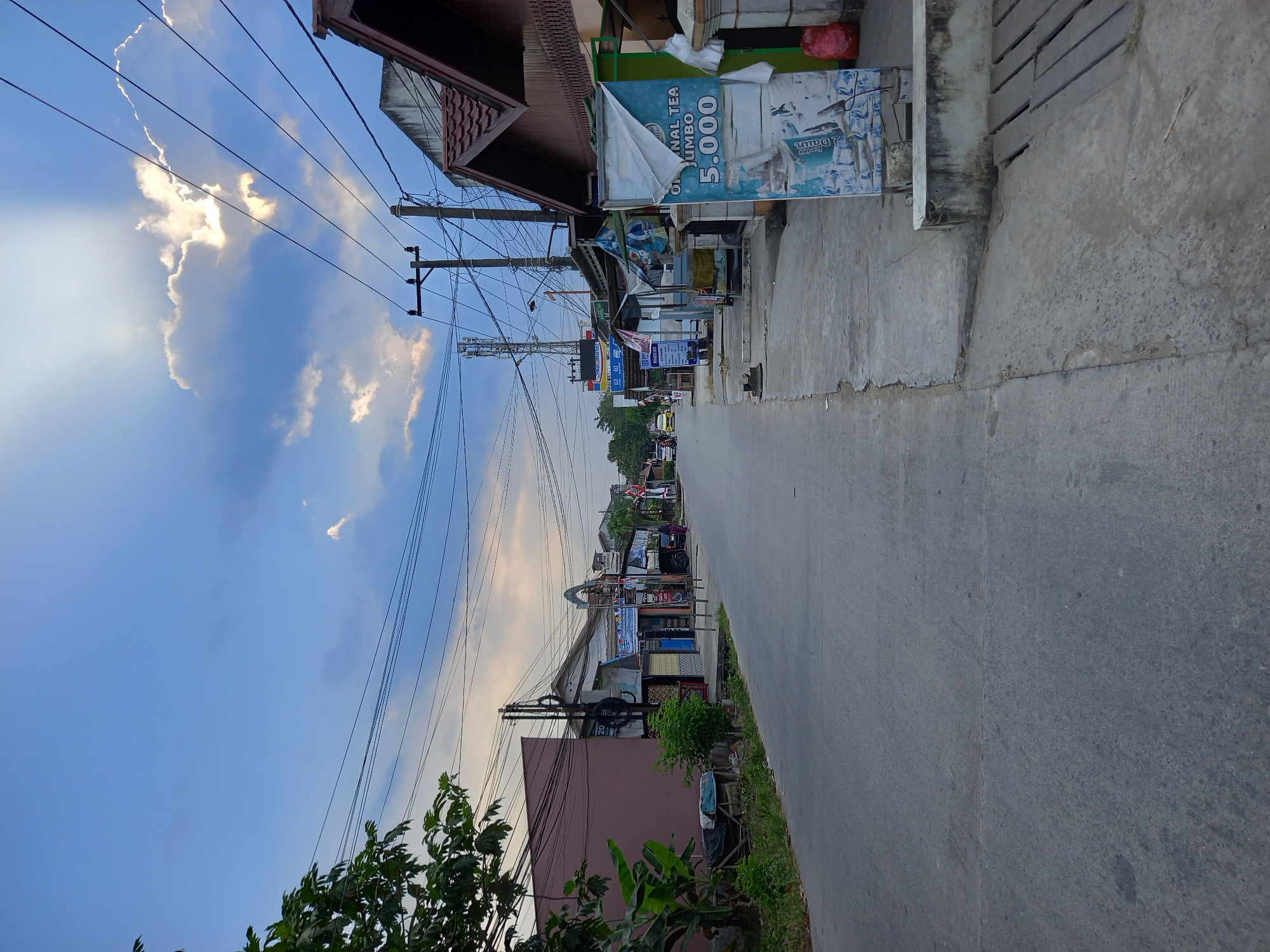 The image depicts a street scene in the city of Samarinda, Indonesia, likely during the late afternoon given the sunlight filtering through the clouds. The street is paved with asphalt and shows signs of wear. On the right side of the street, there is a sidewalk constructed of concrete and wood. Along the sidewalk, various small shops or stalls are visible, some with awnings providing shade. A prominent sign displays "ORIGINAL TEA JUMBO 5.000". Further down the street, there are more buildings and shops, including one with a "Indomaret" logo, suggesting a convenience store. Utility poles with numerous electrical wires are present throughout the scene. In the distance, a telecommunications tower can be seen. On the left side of the street, there are trees and vegetation, and a building with a muted brown facade. The sky is blue with scattered clouds, and shafts of sunlight are breaking through. A few vehicles, including yellow cars and a motorcycle, are present on the street in the distance. A single individual, wearing dark clothing, is walking away from the camera on the sidewalk. A red plastic bag is visible next to the building on the far right. The overall impression is of a typical commercial street in a Southeast Asian city.
