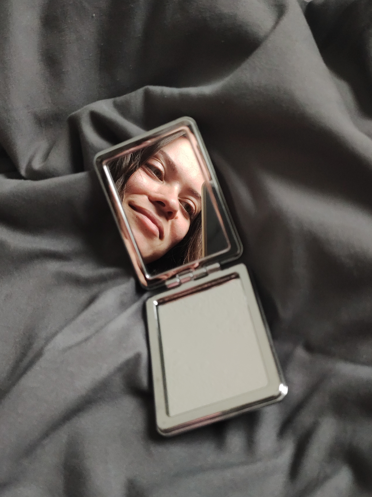The image shows a compact mirror on a gray, wrinkled fabric surface, likely a bedsheet or similar textile. The mirror is open, with the top half angled upwards, reflecting a person's face. The person has fair skin, dark hair, and is looking into the mirror with a slight smile. The bottom half of the mirror is empty, showing its reflective surface. There are no other identifiable objects or people in the scene. The image location is unknown and there is no indication of the city of Chaikovsky, Russia.