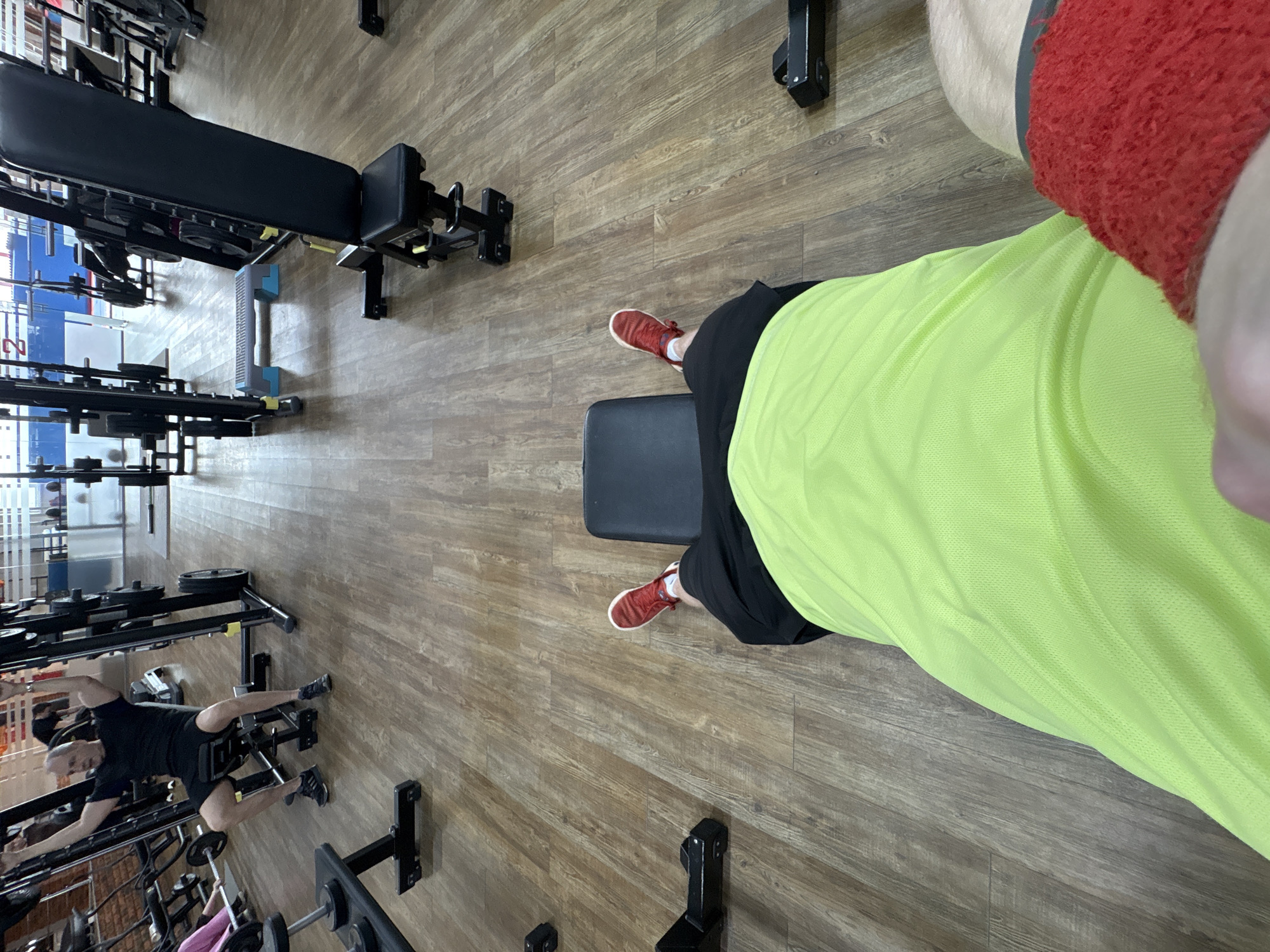 The image shows an interior view of a gym in Chaikovsky, Russia. The gym features wooden flooring and contains various weightlifting machines and benches. A person in a bright green shirt, black shorts, and red sneakers is positioned in the foreground, seemingly seated on a bench. Another person can be seen in the background working out on weightlifting equipment. The gym has a blue and white section in the background near weight racks.