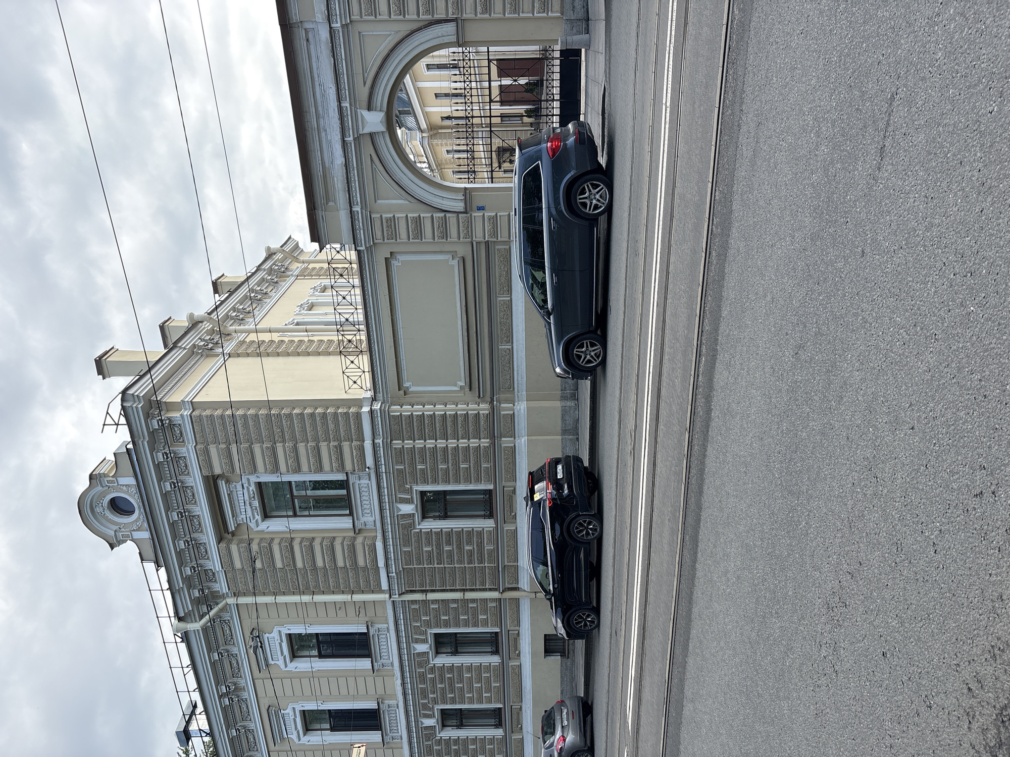 The image depicts a street scene in Saint Petersburg, Russia. A beige building with ornate architectural details stands adjacent to a road with tram lines. Several cars are parked along the street next to the building. The sky is overcast, and power lines are visible in the upper left of the frame.
