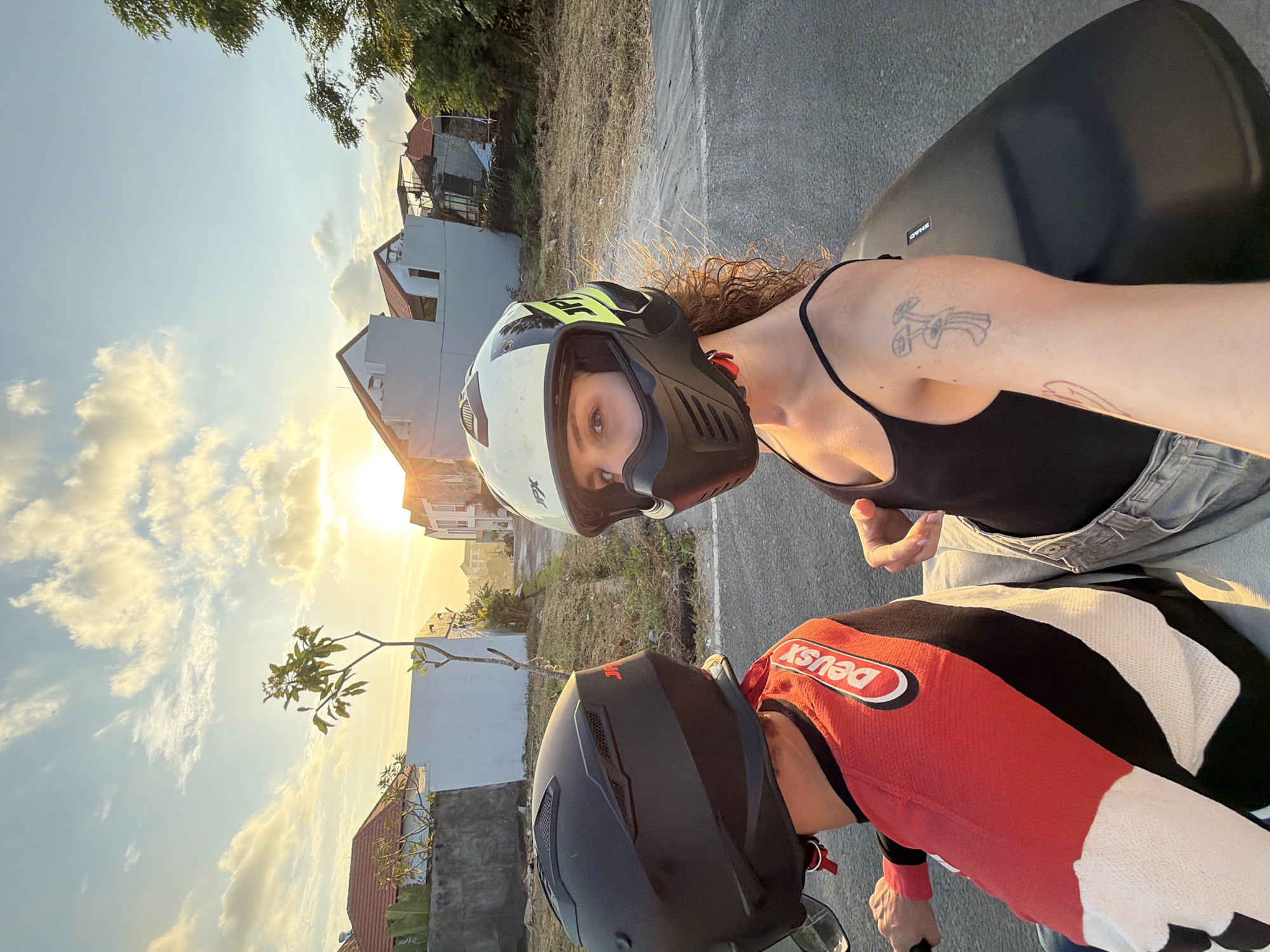 Two individuals are riding a motorcycle in North Kuta, Indonesia. The person in the foreground, wearing a black tank top and denim shorts, is taking a selfie. They have curly brown hair and are wearing a white and yellow helmet with a black face shield. A small tattoo of a mushroom and eye is visible on their right arm. The person in the background is wearing a black helmet and a red, black, and white long-sleeved shirt with the "Deus Ex Machina" logo. The sun is setting, casting a warm glow on the scene, with clouds scattered across the sky. Residential buildings and dry vegetation are visible in the background, suggesting a semi-urban or suburban environment.