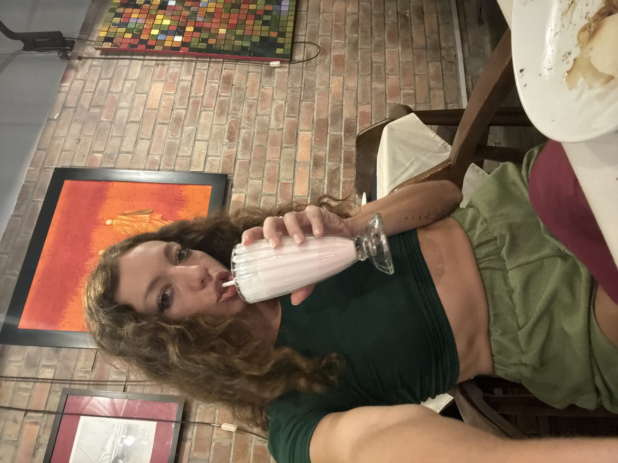 A woman is drinking a milkshake in what appears to be a restaurant. She is wearing a green top and green shorts. There is a plate with food in the upper right corner, and brick walls are visible in the background, suggesting an indoor setting. A painting and a colored-square wall piece hang on the brick wall, indicating a decorated interior. The location is Hà Nội, Vietnam, based on the context and likely locale of the photograph. The woman is holding a milkshake glass, and her pose suggests she is taking a selfie or having someone take a photo of her while drinking.