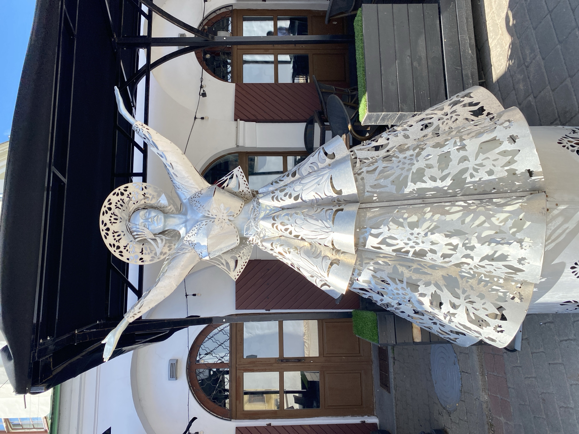 This media file shows a large, metallic sculpture of a woman, likely an angel or a mythical figure, on an outdoor patio. The sculpture is silver and intricately designed with floral and leaf-like cutouts, especially prominent on its flowing dress and headdress. The figure has its arms outstretched, with one hand reaching forward. The sculpture is placed in front of a building with white walls and large wooden doors and windows, some of which are arched. There's a dark, overhead canopy structure above the sculpture, supported by metal beams.
The setting appears to be a public space, possibly a cafe or restaurant patio, given the presence of outdoor seating, including dark wooden tables and chairs visible in the background. A small patch of artificial green turf is also visible. The ground is paved with cobblestones and grey tiles.
The lighting suggests it is daytime, with bright sunlight casting shadows. The sky is visible and appears clear. There is no visible text in the image. The overall mood is artistic and somewhat ethereal due to the sculpture's design. The location context provided is Minsk, Belarus.