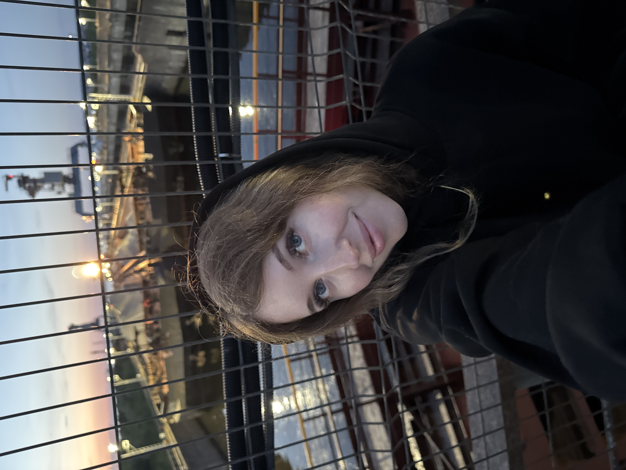 A young woman with light brown hair and blue eyes takes a selfie outdoors in front of a metal fence. She is wearing a black hoodie. Behind her, a large ship is docked in what appears to be a dry dock or canal. The sky is a dusky blue with hints of orange and pink from a sunset. Lights from the surrounding industrial area are visible in the background. The location is the city of Chaikovsky, Russia.