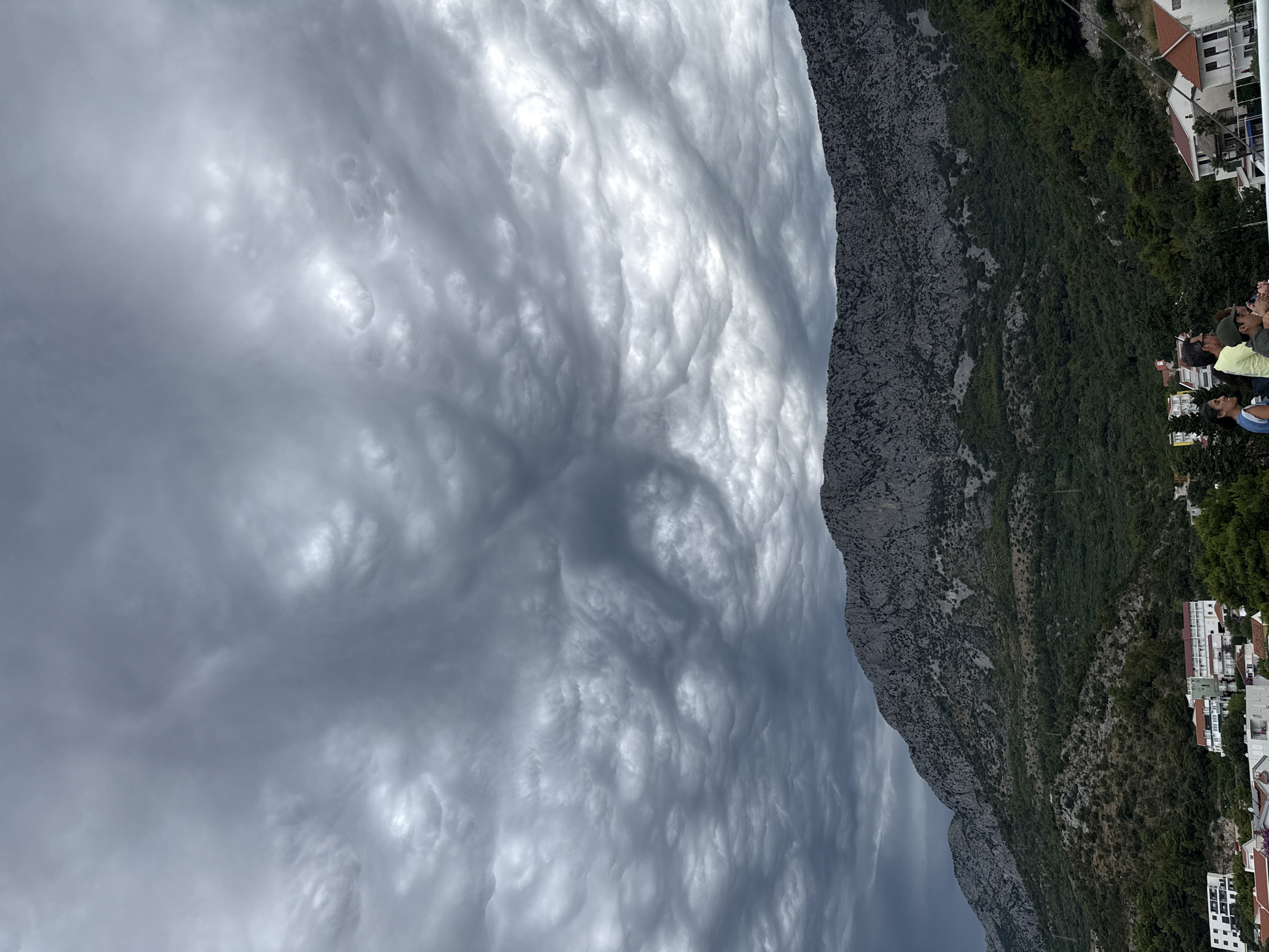 This is a landscape photograph taken from Drvenik, Croatia, showcasing a dramatic sky filled with mammatus clouds over a rugged mountain range. The clouds are dense and gray, with a distinctive undulating pattern, suggesting an impending storm. Below the imposing sky, the steep, rocky slopes of the mountain are covered in patches of green vegetation. Nestled at the base of the mountain are several buildings, indicating a residential or tourist area. In the foreground, partially visible, are a group of people, seemingly observing the sky. Their presence suggests they are either residents or visitors enjoying the view of the unusual cloud formation. The overall atmosphere is one of awe and a touch of foreboding due to the intense cloud display.