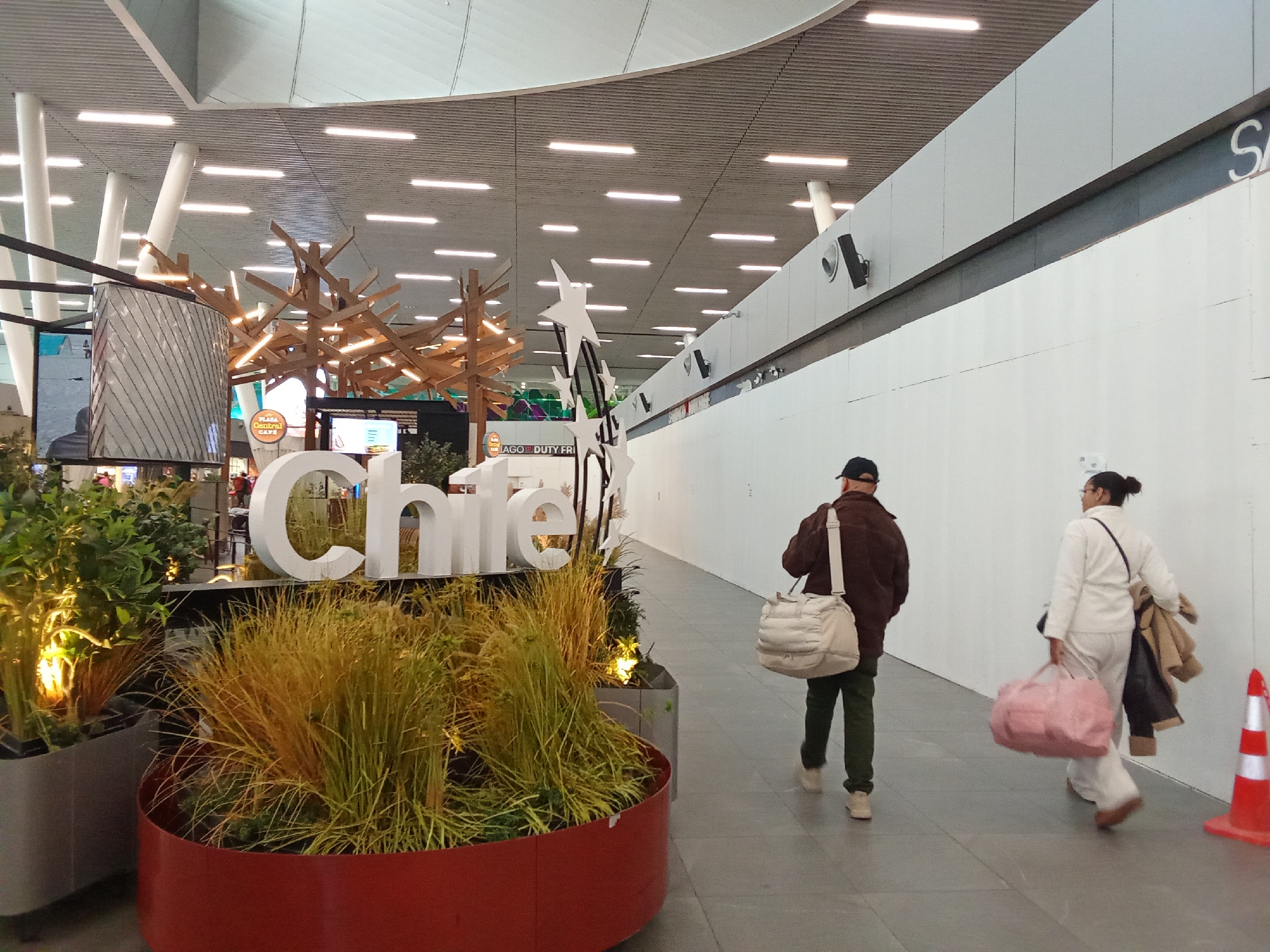 The image shows an indoor scene, likely an airport terminal, based on the large signage, modern architecture, and the presence of travelers with luggage. In the foreground, a large, white, three-dimensional "Chile" sign is prominently displayed amidst lush greenery and ornamental grasses in planters. To the right of the sign, two individuals are walking away from the camera down a hallway. The person on the left is wearing a brown jacket, green pants, and a dark baseball cap, carrying a cream-colored duffel bag over their shoulder. The person on the right is wearing a white outfit, carrying a pink duffel bag and a brown coat draped over their arm. The environment features a high, modern ceiling with integrated lighting, and a large, plain white wall on the right side of the hallway. A bright orange traffic cone is visible near the right edge of the frame. The overall impression is one of a well-lit, contemporary public space. The mention of Pudahuel, Chile, suggests this location is likely within Arturo Merino Benítez International Airport (SCL), which serves Santiago and is located in Pudahuel.