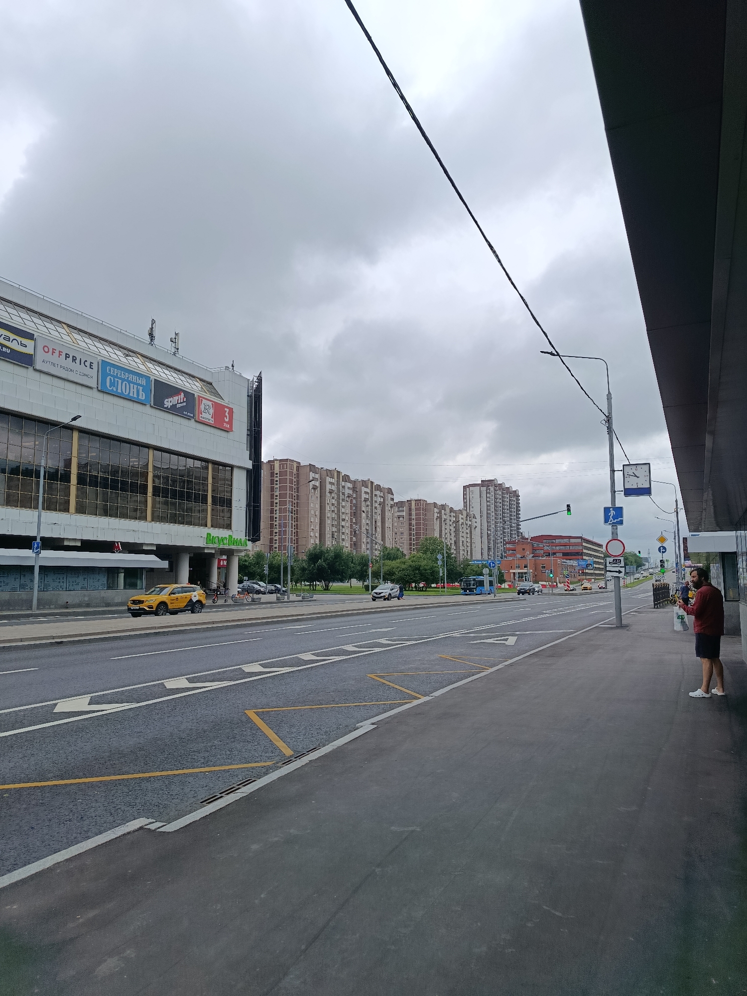The image captures a street scene in Moscow, Russia, under an overcast sky. In the foreground, a paved sidewalk runs along the edge of a wide road with multiple lanes. Traffic signs and a lamppost are visible on the sidewalk. A person wearing a maroon shirt and dark shorts stands on the sidewalk, looking at their phone. Further down the sidewalk, a bus stop shelter is present.
Across the road, a large building with a white facade and dark windows dominates the left side of the frame. Several signs with Russian text and logos are displayed on its upper section, including "OFFPRICE" and "СЕРЕБРЯНЫЙ СЛОНЪ". A yellow taxi is parked in front of this building.
In the background, a row of tall, multi-story apartment buildings, typical of Soviet-era architecture, stretches along the horizon. Trees and greenery are interspersed between the buildings and along the roadside. A blue bus is traveling on the road, and other vehicles are present in the distance. The overall atmosphere suggests a typical urban environment on a cloudy day.