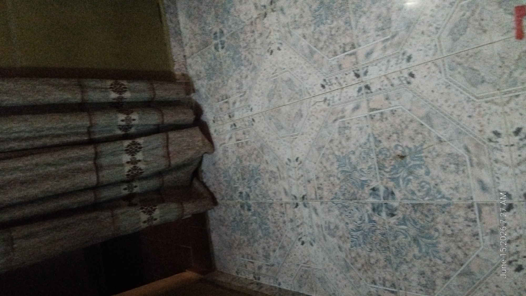 The image depicts a floor tiled with a light-colored pattern. A dark-colored curtain with a repetitive floral design hangs across the top portion of the frame. The curtain is positioned to the left, obscuring a portion of the wall and possibly a doorway. The floor tiles have a somewhat intricate pattern, primarily in shades of light blue and beige, with some darker speckling. There is a timestamp in the bottom left corner indicating "June 15, 2026 7:27 AM". The scene appears to be indoors, likely a room. There is no indication of the specific location within the city of Howayi, Nigeria, as the image does not contain any identifying landmarks or context clues for that specific geographical area.