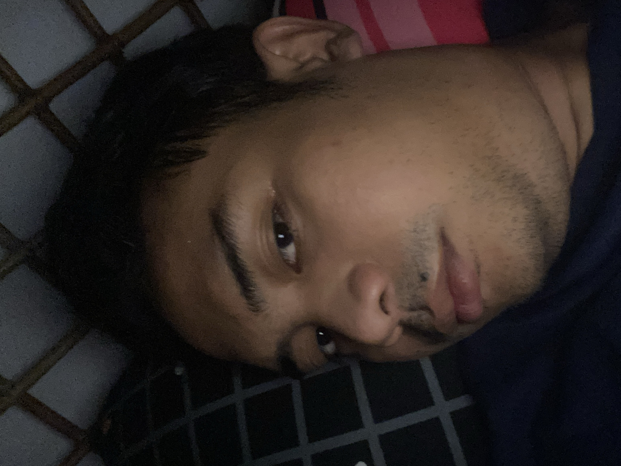 The image shows a close-up, head-and-shoulders view of a man of Southeast Asian descent. He has dark hair, brown eyes, and appears to be lying down. The background includes a dark-colored ceiling with a grid-like pattern, suggesting he may be indoors. A patterned bedsheet or fabric is visible behind his right shoulder. The man is looking directly at the camera, with his mouth slightly open. The lighting is dim, casting shadows on his face. The image does not provide any specific visual cues to indicate the location is in Dukuh, Indonesia.