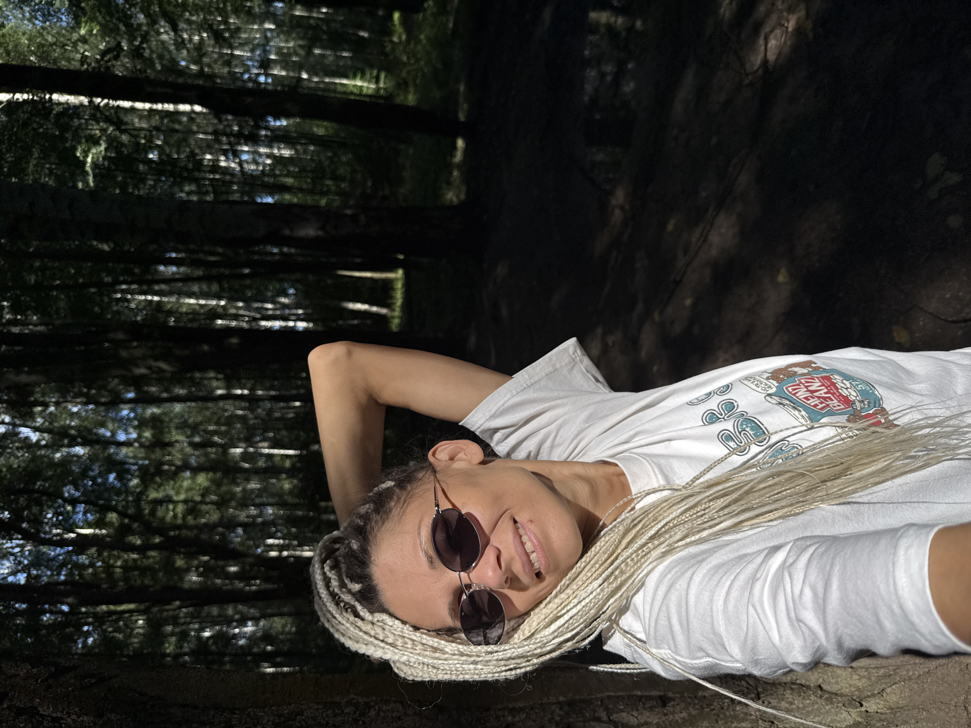 A medium shot captures a fair-skinned woman with long, blonde braids posing for a selfie in a sun-dappled forest. She is leaning against a tree trunk on the left, her right arm raised behind her head, and her left hand is not visible. The woman is wearing a white t-shirt with a graphic print. She has on oval, dark-lensed sunglasses and is smiling at the camera, her teeth visible. The background is a dense forest with tall, slender trees and dappled sunlight filtering through the leaves, creating a chiaroscuro effect. The ground is covered in dark soil and fallen leaves. The lighting suggests it is daytime, possibly late afternoon, with the sun casting long shadows. The image appears to be taken outdoors in a natural setting. The location is identified as Rusanovo, Russia.