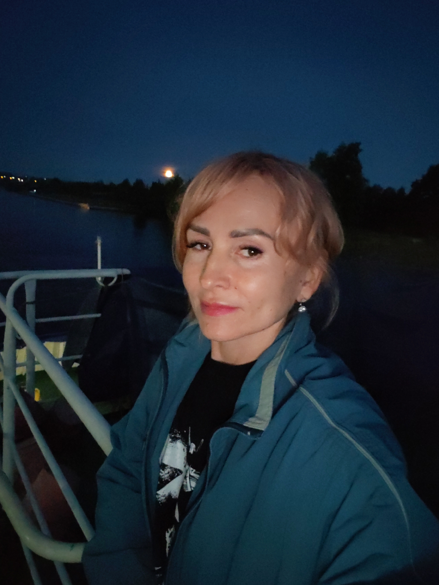 A woman with blonde hair and a blue jacket is taking a selfie at dusk. She is on what appears to be a boat or dock, with a railing in the foreground. In the background, there is a body of water and trees, with a faint light on the horizon. The sky is dark blue, indicating it is nighttime.