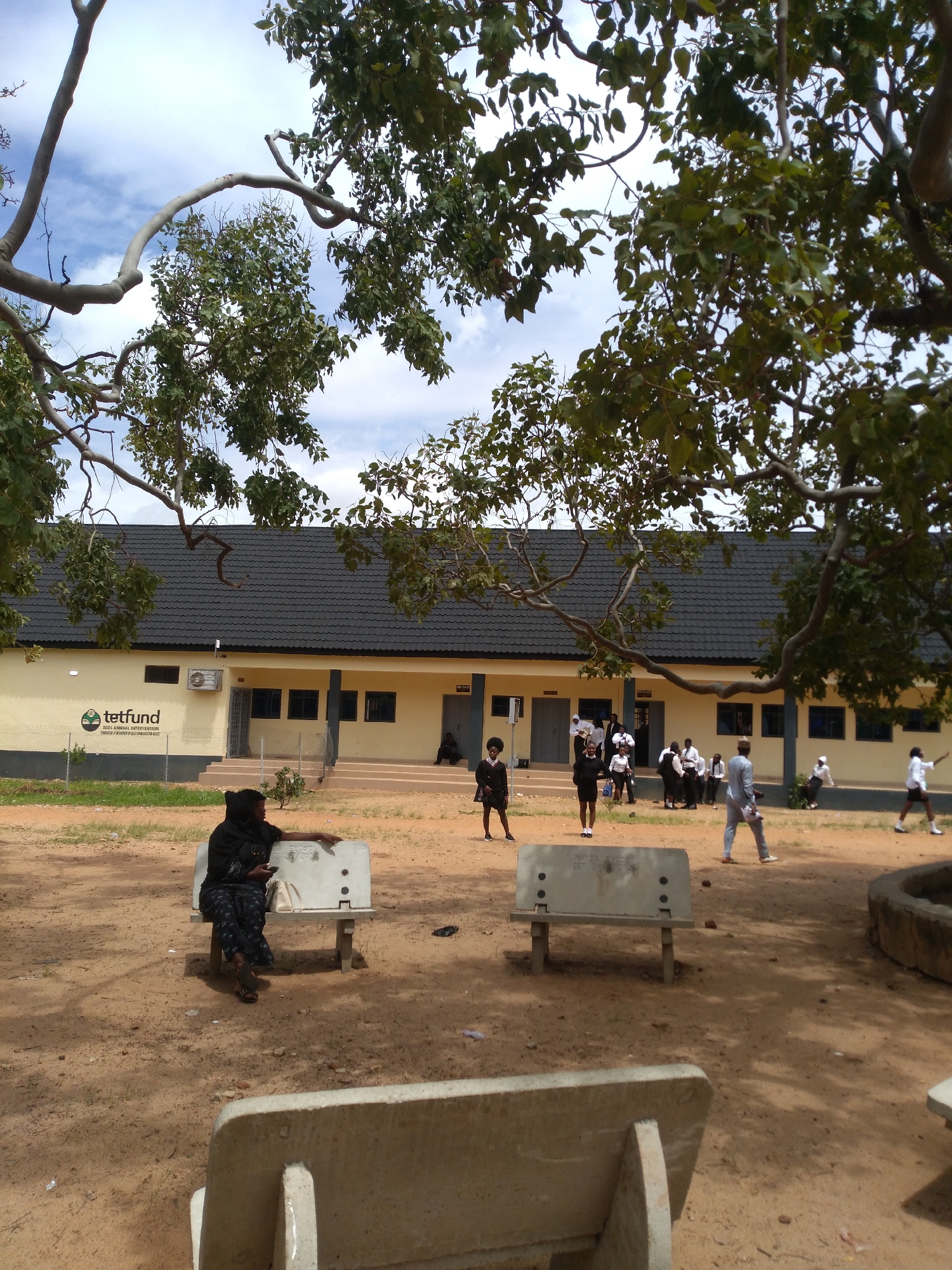 The image displays an outdoor scene in the city of Jos, Nigeria, featuring a light yellow, single-story building with a dark grey tiled roof, multiple individuals, concrete benches, and trees under a partially cloudy sky.
The building occupies the mid-ground, presenting a pale yellow facade with several dark-framed windows and two prominent entrances, each featuring a set of steps. An air conditioning unit is mounted on the left side of the building. A white logo for "tetfund" and the accompanying text "2021 ANNUAL INTERVENTION" are visible on the building's left wall.
In the foreground, a woman in a black head covering and dark, patterned attire is seated on a light-colored concrete bench, facing left. She holds a mobile phone in her right hand, which rests on her lap, and her left arm is extended over the backrest of the bench. A white bag is positioned next to her on the bench. Inscriptions reading "EXCO 20/03/23" are visible on the backrest of this bench. Closer to the viewer, the backrest of another concrete bench is visible. A third concrete bench is partially visible to the right, bearing the inscription "HATS S.C.C.E.L. SBT 2016".
The ground consists primarily of exposed, reddish-brown soil or sand. Sparse green vegetation is visible in the background, beyond a low wire fence. Numerous individuals, many dressed in white shirts and dark lower garments, are gathered and moving in the mid-ground near the building. One person in a dark suit stands to the left, while another in a light-colored suit walks towards the right.
Large trees with green foliage frame the upper and side portions of the image, with their branches extending across the sky. The sky above is blue with scattered white clouds.