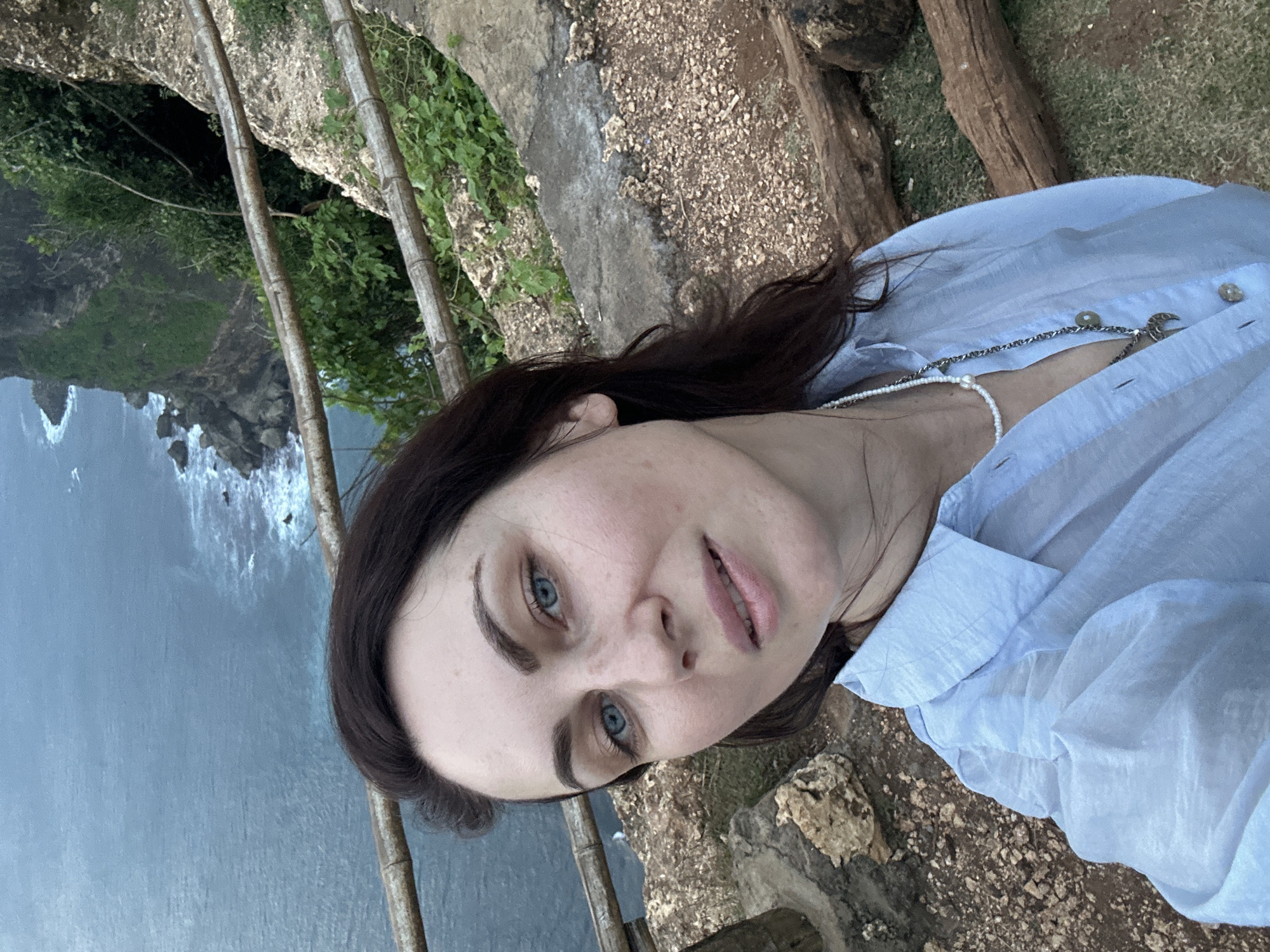 A selfie of a woman is taken from a high vantage point overlooking the ocean in Kuta Selatan, Indonesia. The woman has dark brown hair and blue eyes and wears a light blue button-down shirt and two necklaces, one with beads and one with a pendant. In the background, cliffs drop off to the water, and waves crash against the rocks. A crude bamboo fence is visible between the woman and the cliff edge. The foreground shows rocky ground covered with sparse vegetation.