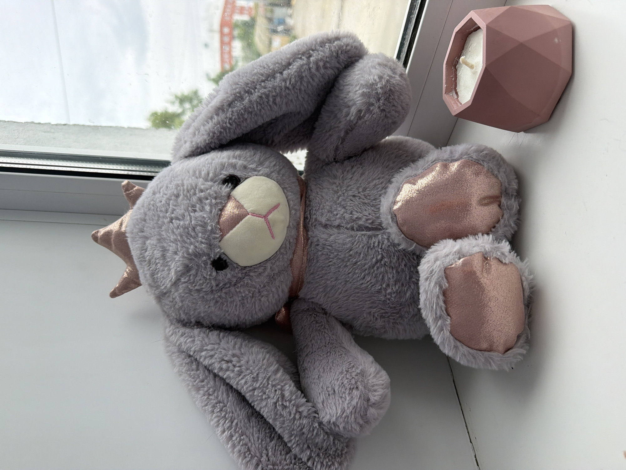 A plush gray bunny wearing a pink crown and collar sits on a white windowsill. A pink geometric candle holder with a white candle sits to the right of the bunny. Through the window, a cloudy sky and some buildings are visible. The scene is indoors, possibly in an apartment in Yuzhno-Sakhalinsk, Russia.