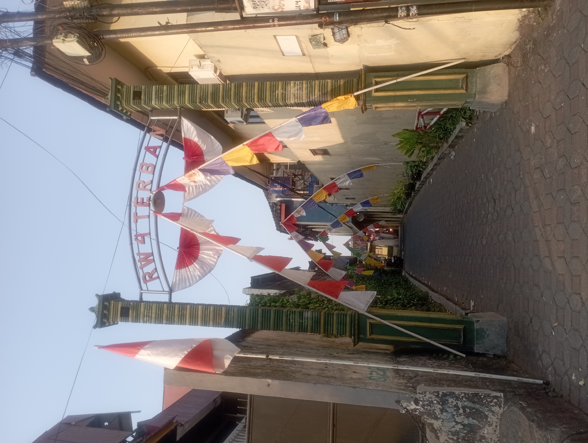 The image depicts a narrow, paved alleyway in Yogyakarta, Indonesia, decorated with Indonesian flags and pennants. An archway at the entrance to the alley bears the text "RW 4 TERBAN," indicating this is the entrance to neighborhood number 4 of the Terban area. The flags are predominantly red and white, with some pennants featuring multiple colors such as yellow, blue, and white. The buildings flanking the alley are two-story structures with light-colored walls. An air conditioning unit is visible on one of the building walls. Overhead wires and electrical boxes are present. The alley leads deeper into a residential area, where a motorcycle is parked. The scene appears to be set during daylight hours, with the sun casting shadows on the ground and walls.