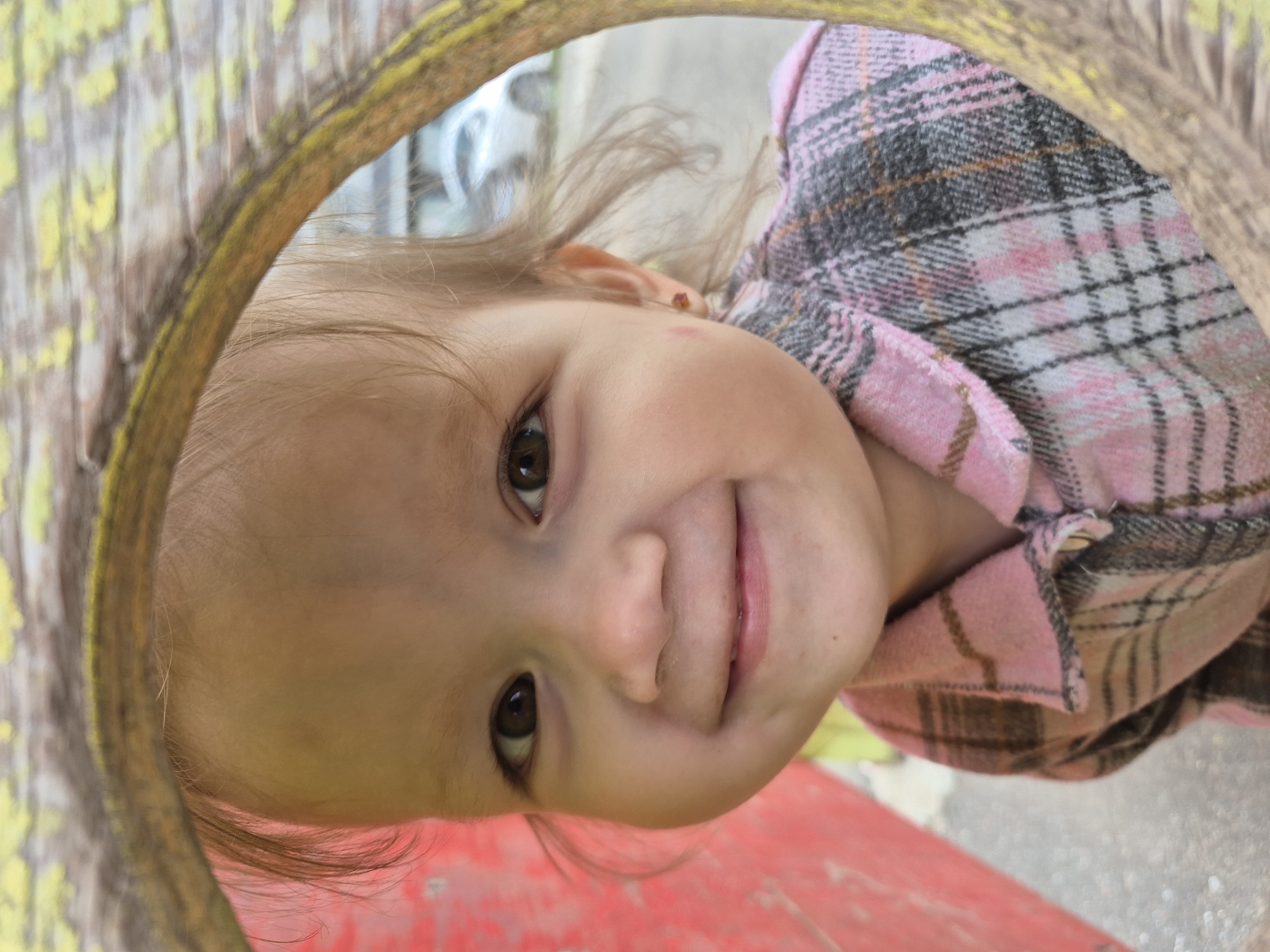 A close-up, angled shot shows a young child peeking through a circular opening, which appears to be a tire or tunnel. The child, with light brown hair and wide brown eyes, is looking directly at the camera with a slight smile. They are wearing a pink and gray plaid shirt. The opening frames the child's face and upper body, with the rest of the scene blurred in the background. The background suggests an outdoor setting, possibly a playground, with hints of colorful surfaces and muted outdoor elements. The lighting appears to be natural daylight, possibly overcast given the soft shadows. The child's expression is one of curiosity and openness, a common emotion for a child engaged in play or exploring. There is no visible text in the image. The setting appears to be a casual, outdoor environment.